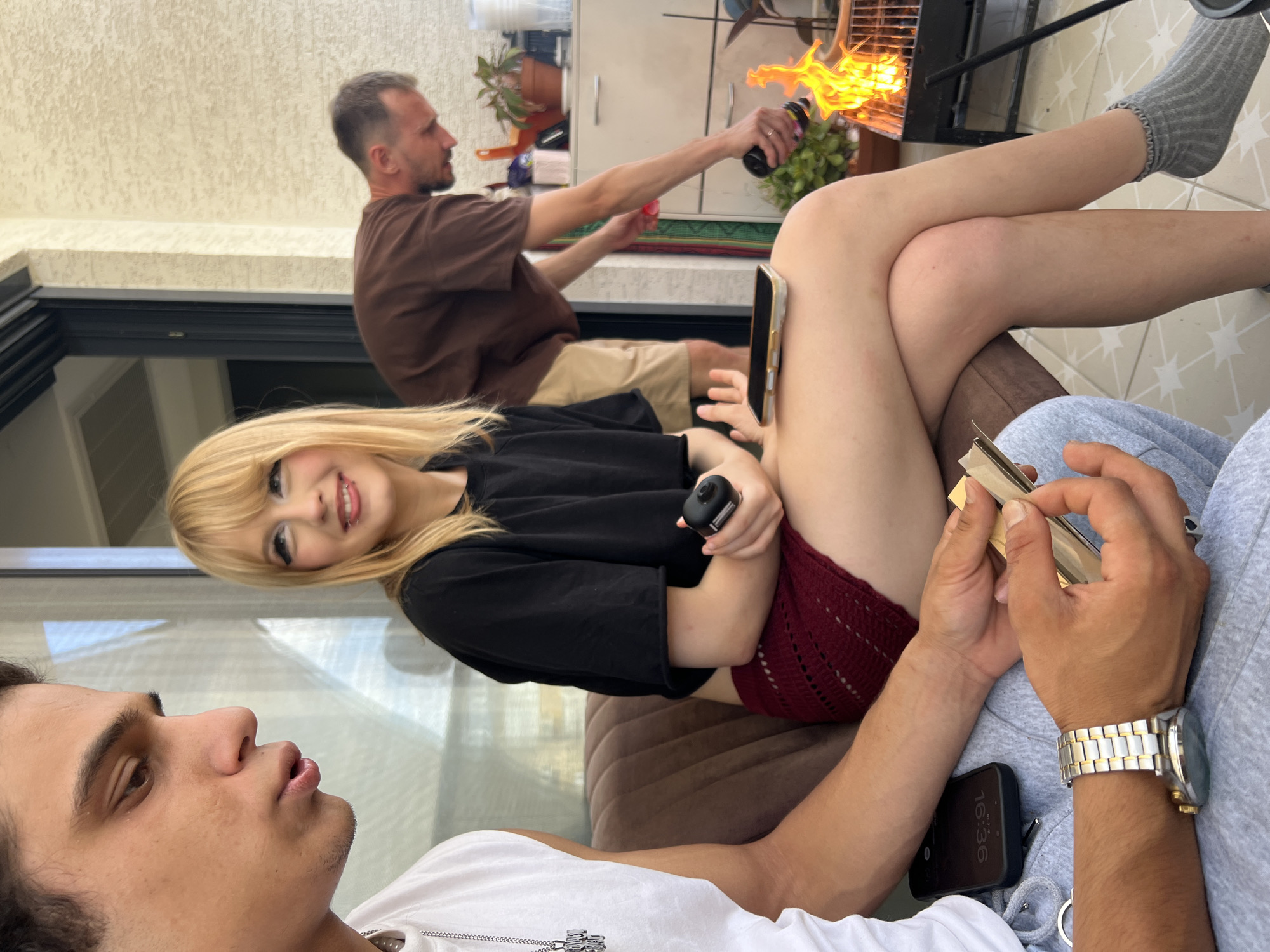 In Kiryat Bialik, Israel, on a balcony, three individuals are present. In the foreground, a young man wearing a white t-shirt and a silver watch sits on a brown couch, holding a rolling paper and a tobacco pouch. To his right, a young woman with blonde hair and heavy eye makeup sits on the same couch, wearing a black crop top and a red skirt. She holds a small black object in her right hand and a smartphone on her lap. In the background, a man in a brown t-shirt and beige shorts is tending to a barbecue, holding a lighter. The balcony floor is tiled, and a railing is visible. Various items, including plants, a grill, and cabinetry, are present on the balcony. The overall setting suggests a casual outdoor gathering.