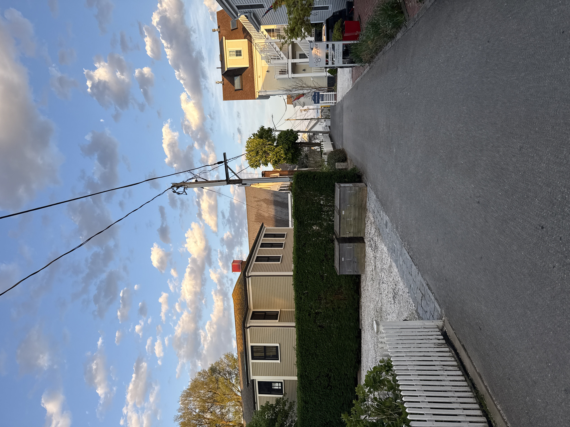 The image depicts a street scene in Provincetown, Massachusetts, USA, characterized by a narrow asphalt road flanked by buildings and vegetation. The sky is filled with scattered cumulus clouds, illuminated by the late afternoon sun. On the left side of the road, a residential building with light-colored siding and dark-framed windows is partially obscured by a tall, manicured hedge. Two wooden planters are visible adjacent to the hedge. A white picket fence runs along the edge of the road, bordered by foliage. Overhead, power lines stretch across the sky. The right side of the road features multi-story buildings with white exteriors and architectural details like balconies and staircases. A street sign is visible, partially obscured by foliage. The overall impression is that of a quiet residential street during the late afternoon.