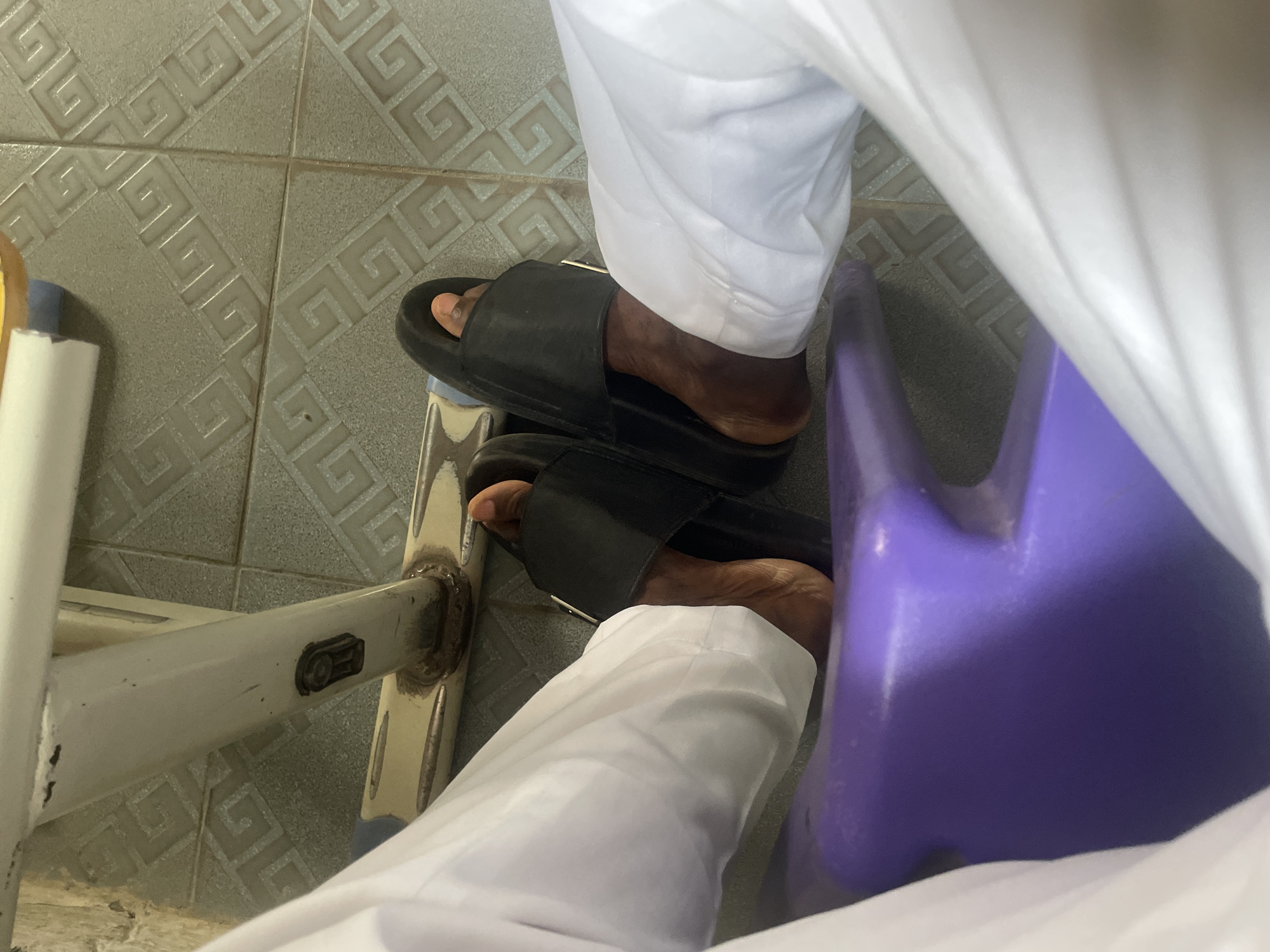 The image shows a close-up view of a person's legs and feet wearing white clothing and black slides, resting on a purple plastic object, possibly a chair or stool. The background consists of tiled flooring with a repeating geometric pattern. The scene appears to be indoors, with the lighting suggesting it might be daytime. The context of Abuja, Nigeria, cannot be determined from the visual information provided in the image.
