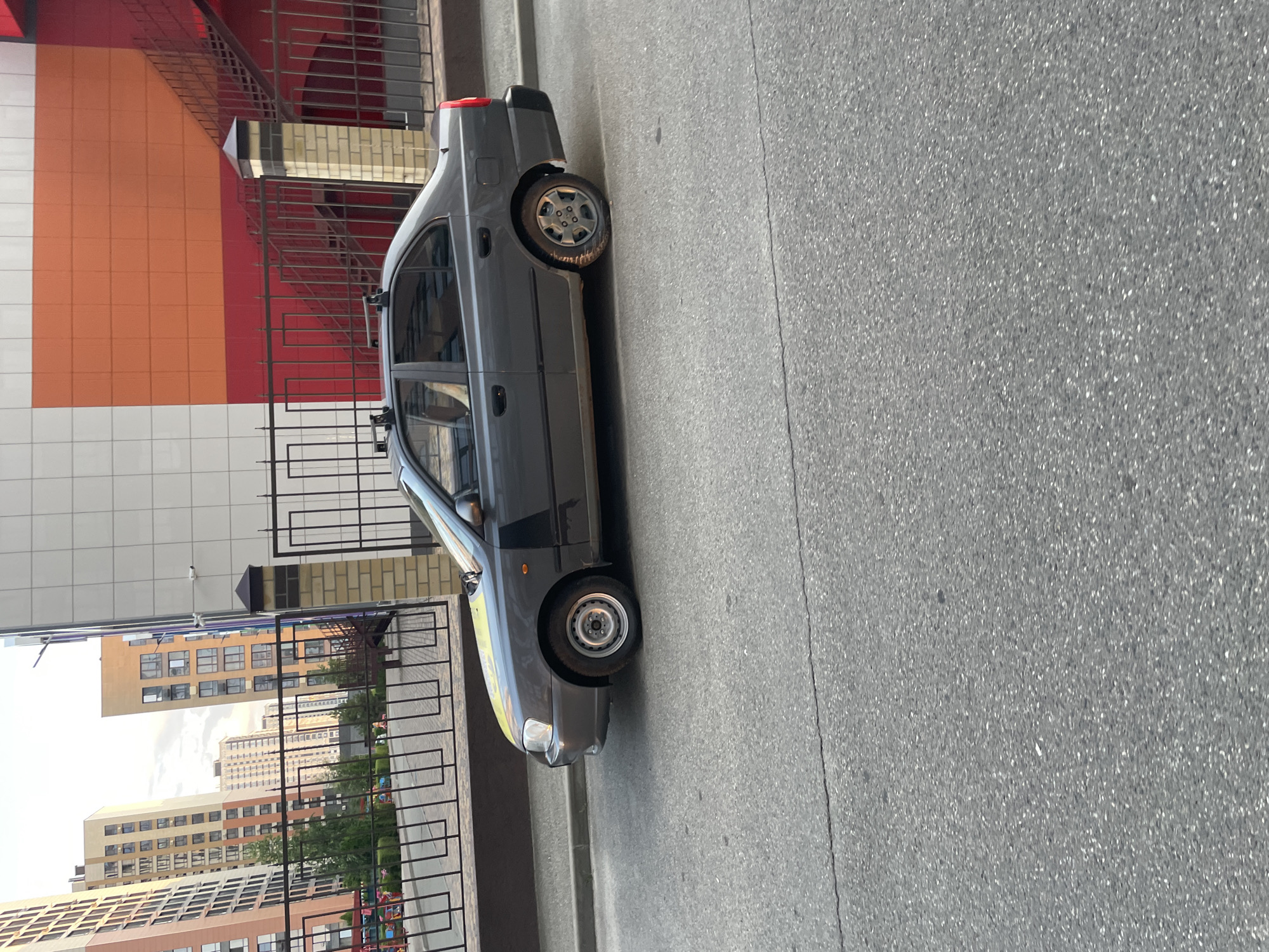 A dark grey sedan is parked on an asphalt road, its right rear tire appears to be deflated. The car has a roof rack. Behind the car is a black metal fence, a brick pillar, and a building with orange and red colored panels. In the background, tall residential buildings are visible. The scene appears to be in an urban environment.