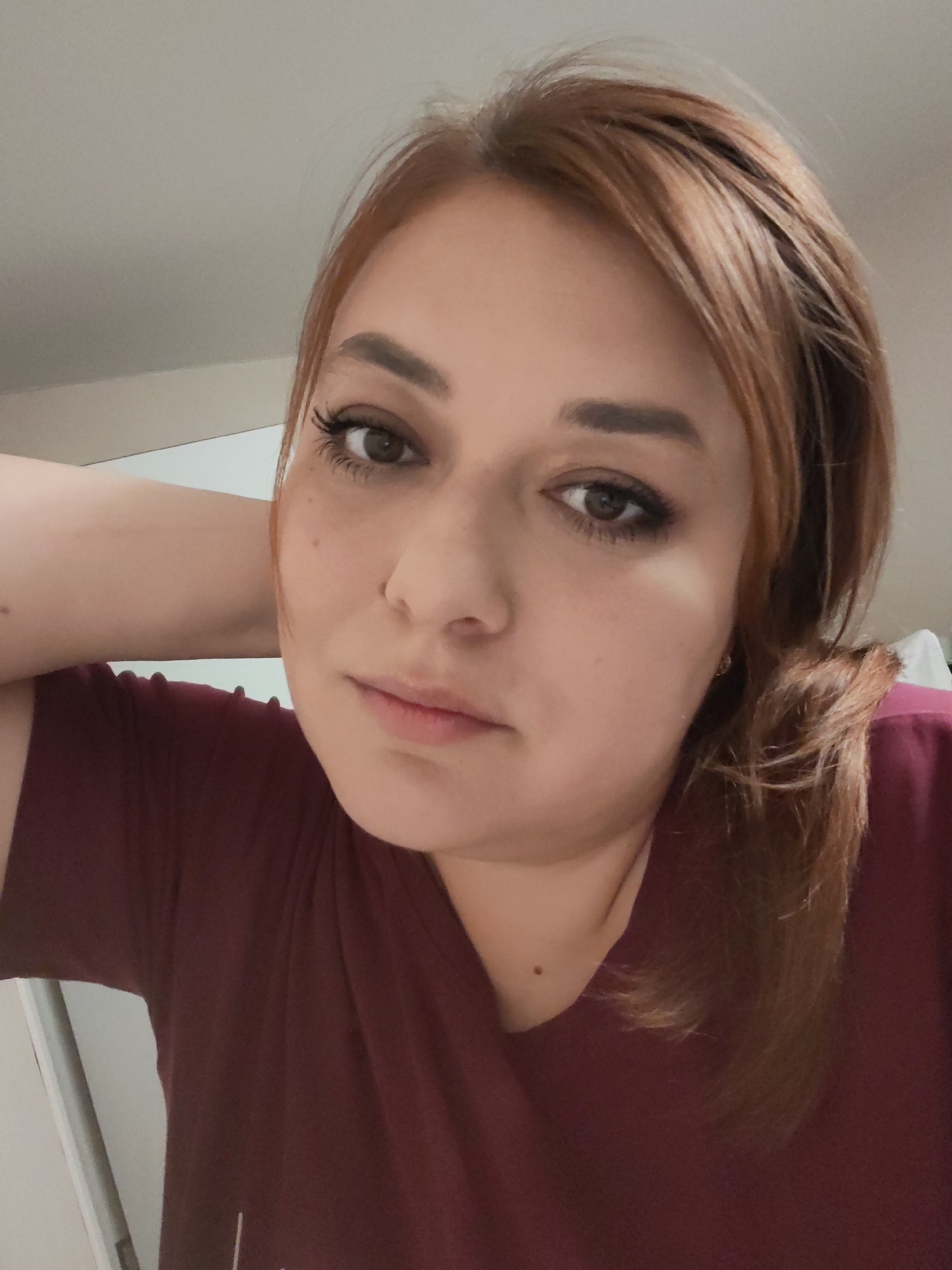 A woman with auburn hair and brown eyes poses for a selfie. She is wearing a maroon t-shirt and has her left hand behind her head. The background is a plain white wall and part of a white door frame. It is not possible to determine if the location is in the city of Podolsk, Russia from the image.