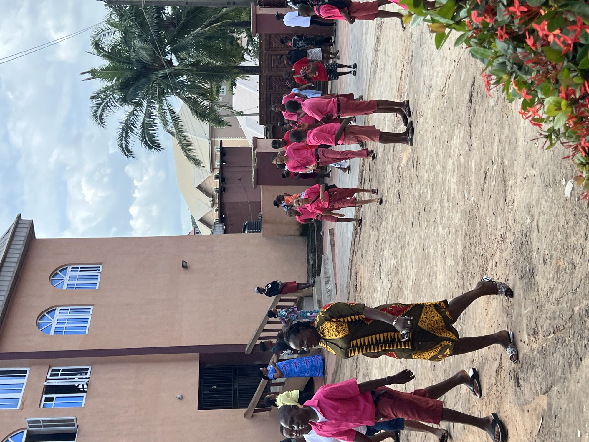 In Nnewi, Nigeria, a group of people, predominantly young individuals dressed in matching pink shirts and red shorts, are gathered outdoors. The scene appears to be in front of a light brown, multi-story building with arched windows. A woman in a vibrantly patterned dress is walking towards the foreground, while other individuals are seen in the background, some standing and others in motion. The ground is a dirt or gravel surface, and lush green foliage with red flowers is visible in the lower right corner of the frame. A large palm tree is also present in the background, suggesting a warm climate. The overall atmosphere suggests a gathering or event, possibly related to a school or community activity.