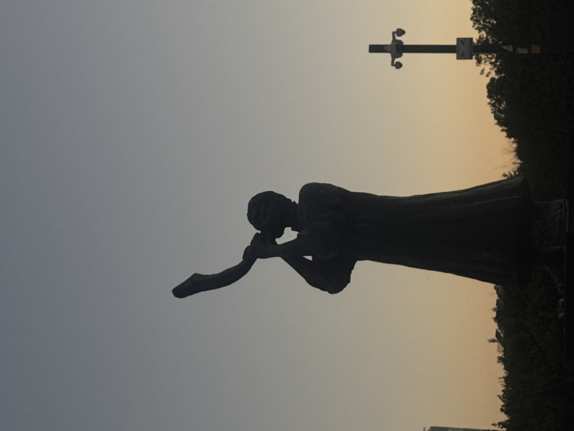 This is a silhouette of a statue against a dusky sky. The statue depicts a person in a long robe, blowing into a shofar, a ram's horn. To the right of the statue, a lamppost with cameras is visible. The background shows a dark line of trees and a faint silhouette of buildings. The statue is located in Ramat Gan, Israel, at the Gush Dan Promenade. The sky displays a gradient from light blue at the top to a pale orange near the horizon, suggesting either dawn or dusk.