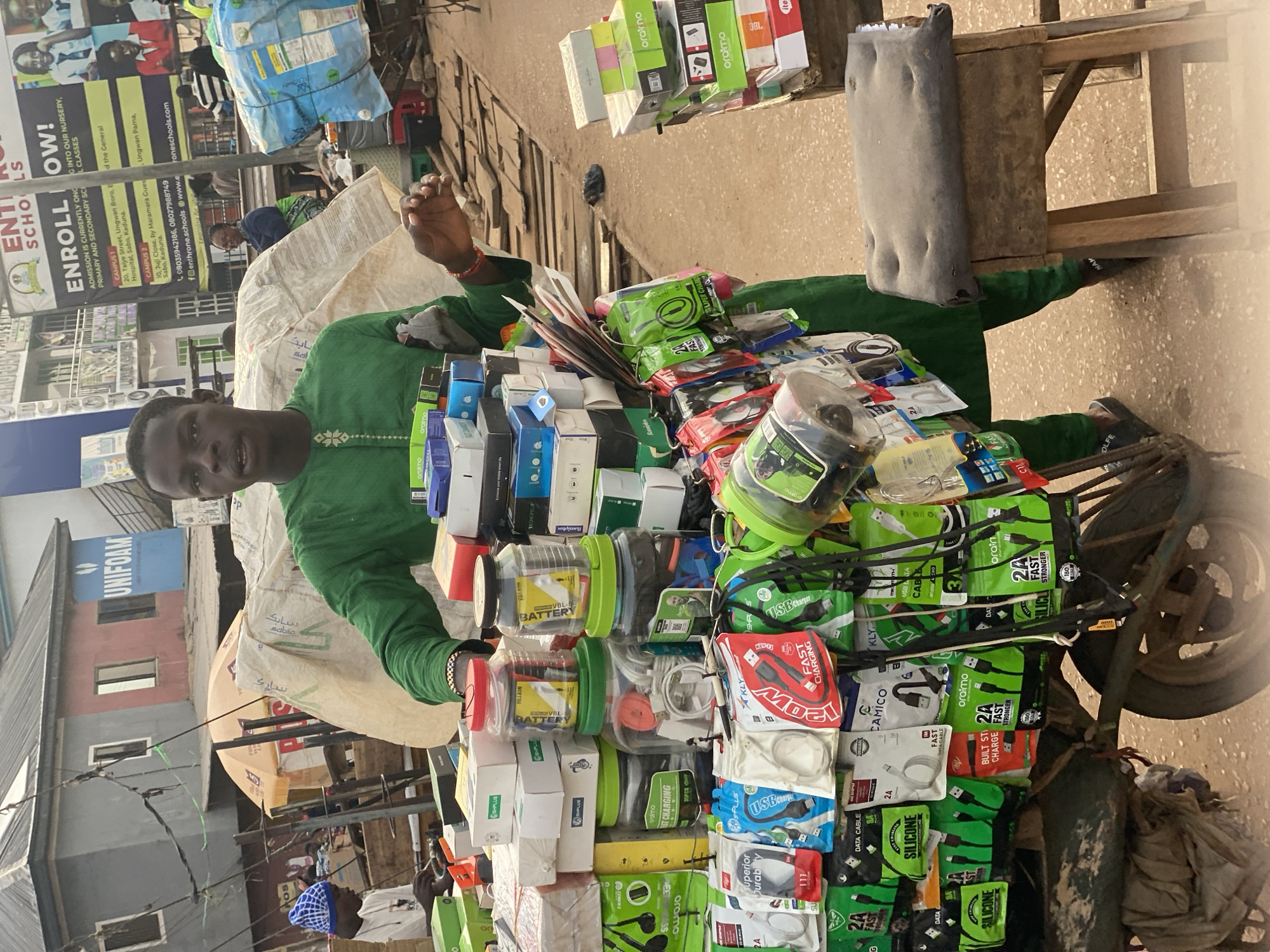 A young Black male, appearing to be in his early twenties, stands behind a mobile cart filled with electronic accessories. He is wearing a bright green traditional outfit. The cart is laden with various items including boxed chargers, cables, headphones, and power banks. Two large transparent jars containing what appear to be batteries and charging cables are prominently displayed. The man is holding a white pen in his right hand, and his left hand is resting on the cart. He is looking directly at the camera with a neutral expression.
The background reveals a busy street scene in Sabon Tasha, Nigeria. Buildings with corrugated iron roofs and signs are visible. One large billboard in the background advertises "ENTHRONE SCHOOLS" with enrollment information. Other individuals are sparsely visible in the distance, including a person wearing a blue and white patterned hat. The ground is dusty and appears to be a dirt road. A wooden stool with a worn, dark seat is placed to the right of the cart. The overall atmosphere suggests a bustling market environment.