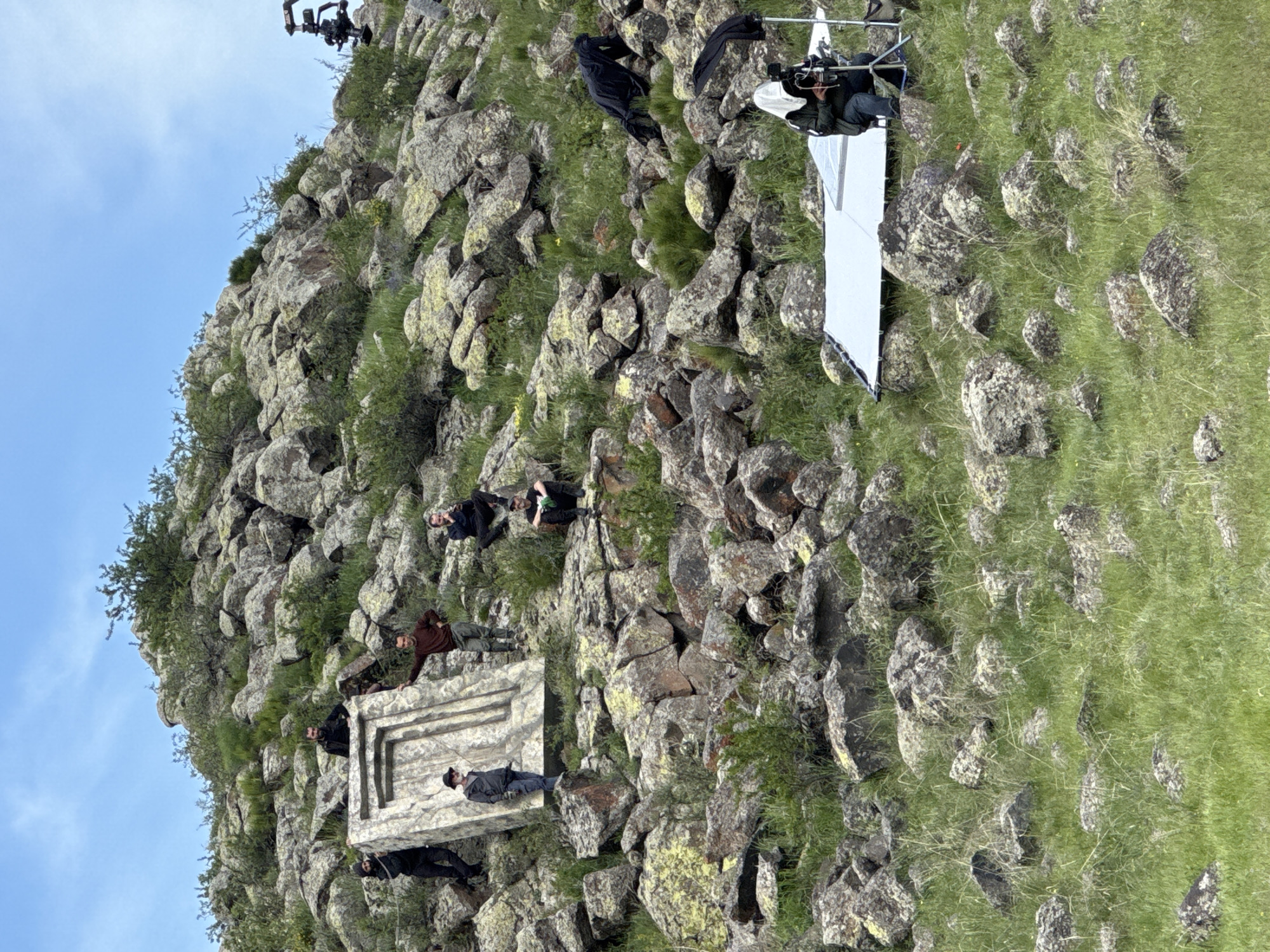 The image depicts a film set on a rocky hillside, likely in a rural area. People are present, some appearing to be crew members operating cameras and lighting equipment, while others seem to be actors positioned near a rectangular stone structure with a raised border. The structure is sitting on the hillside. The environment consists of rocks, green vegetation, and a clear blue sky. The location is Tlik, Armenia. The action appears to be a film shoot.
