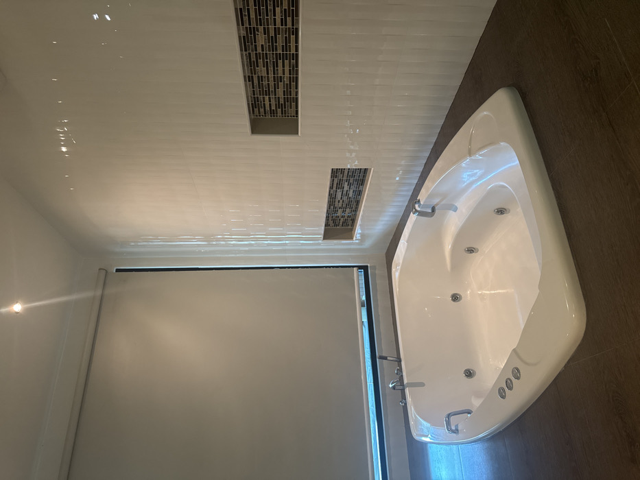 This image depicts a clean, unoccupied modern bathroom located in Ban Ko Li Pe, Thailand. The central subject is a large, white, rectangular jacuzzi bathtub with rounded corners, positioned on a dark brown floor, possibly laminate or large tiles. The bathtub features multiple visible jets, chrome faucets with a shower wand on its left edge, and two chrome grab handles on its rim. Three circular white control buttons are present on the tub's front-facing edge.
The walls are covered with white vertically-textured tiles. Integrated into the tiled wall on the right are two recessed niches, each backed with a mosaic pattern of small rectangular tiles in shades of brown, black, and white. To the left, a large window is covered by a light-colored (beige/off-white) roller blind, obscuring the view outside. The white ceiling contains a single illuminated recessed spotlight in the upper left corner, casting light into the room. No individuals or personal items are present in the scene.