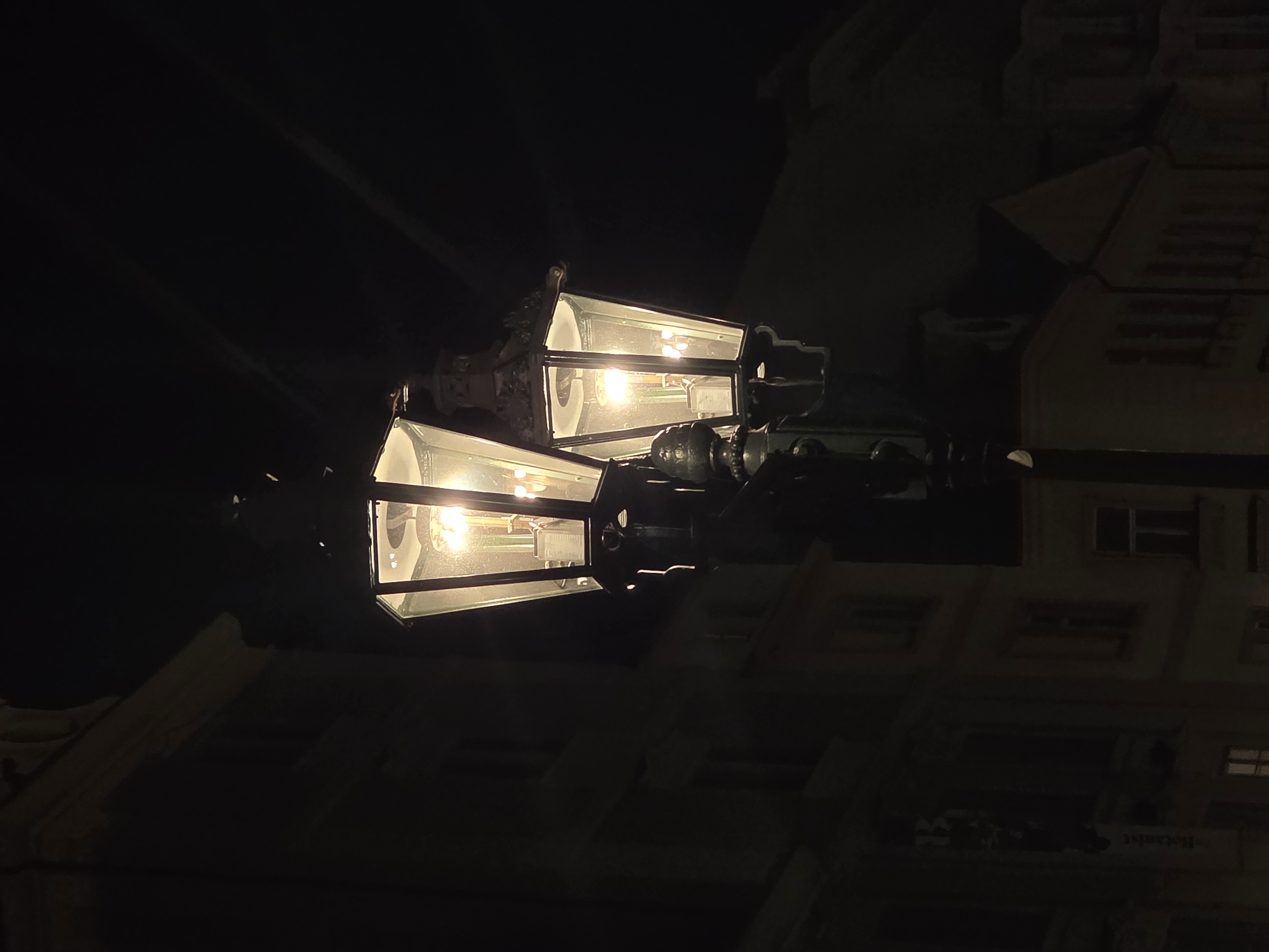The image displays a tall, dark, ornate street lamp featuring two hexagonal or octagonal lanterns, both illuminated, against a dark night sky. The lamppost, likely cast iron, exhibits decorative elements including finials and scrollwork. Each lantern encases multiple incandescent-style bulbs that emit a warm, yellow light through its glass panels. The upper portion of the scene is dominated by a deep black, starless sky. In the background, dark silhouettes of multi-story urban buildings with discernible windows are visible on both sides of the lamppost, suggesting an unknown city at night. Architectural features such as ledges, frames, and rooflines are faintly visible on these structures, which are primarily in shadow, receiving minimal ambient light from the street lamp. No people or active elements are present in the static scene.