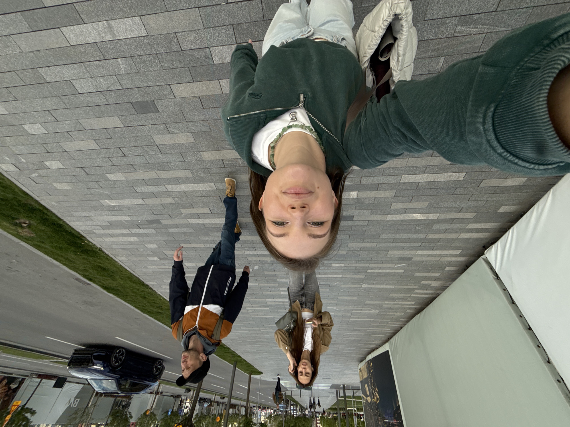 The image shows a selfie taken from an unusual perspective, appearing as though the ground is oriented vertically. In the foreground, a young woman in a green jacket and white shirt is taking the selfie. Behind her, a man in a black and brown jacket walks on the left side of the image, with a car visible in the background on a street. Another woman in a brown jacket walks towards the right, seemingly mirrored. The backdrop includes a street scene with trees, buildings, and pedestrian elements, seemingly inverted due to the camera angle. A white structure is present on the right side. Based on these elements, the location is likely Belgrade, Serbia, because the location on the street is also mirrored.