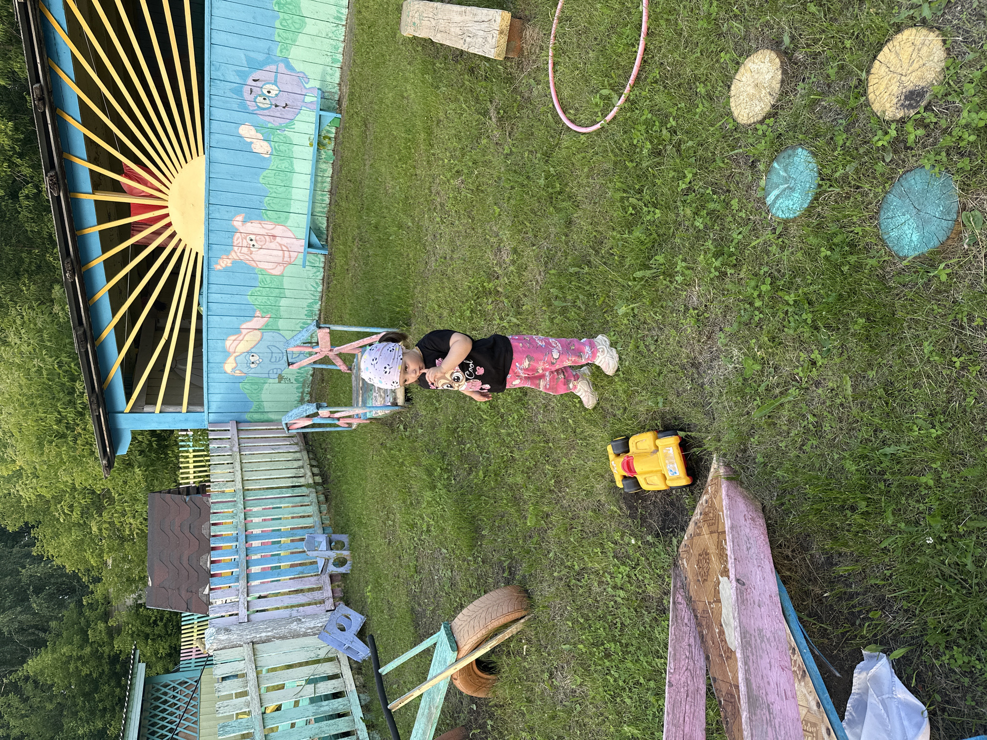 A young child, wearing a bandana and a black t-shirt with a graphic print, stands in a grassy area with toys and playground equipment. The child is holding an object to their mouth. In the background, there is a colorful wooden fence with cartoon characters painted on it, and a building with a sun design. The environment appears to be a playground in a rural or suburban setting, possibly in Yaroslavka, Russia, given the context. There is a yellow toy car on the ground near the child, and other playground items such as a wooden bench, stepping stones made from logs, and a pink hula hoop are visible. The grass is green and somewhat uneven, with patches of clover and weeds. The lighting suggests it is daytime, possibly late afternoon or early evening.