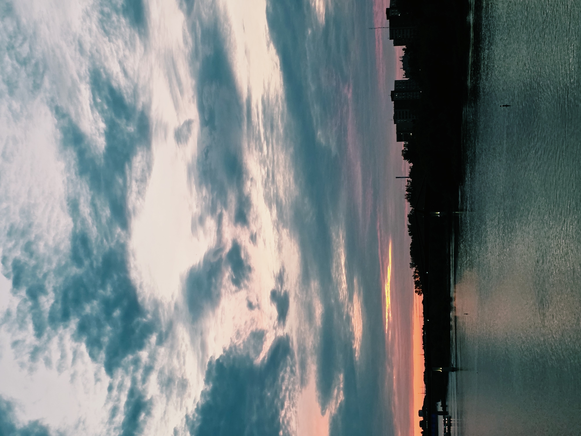 The image depicts a view of the Vistula River in Warsaw, Poland, at dusk. The sky is filled with dramatic, textured clouds in shades of teal, pink, and pale lavender, with a bright band of golden light on the horizon. Silhouetted against the sky are apartment buildings and trees along the riverbank. A bridge is visible in the distance on the left, with lights indicating traffic. The water of the Vistula is calm and reflects the colors of the sky. A single buoy stands in the middle of the river. The overall impression is one of a serene, urban landscape at twilight.