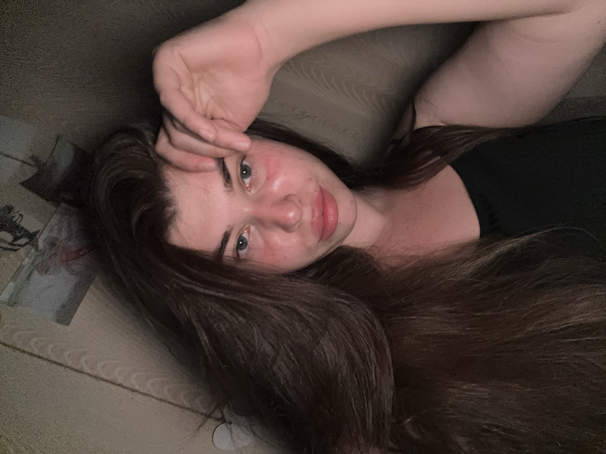 A young woman with long, dark brown hair and blue eyes is pictured in a selfie. She is wearing a black tank top. Her face is slightly flushed, and her hand is resting on her forehead. The background appears to be a dimly lit room with a textured wall and a picture or poster hanging above her. The image does not provide any information about her location in the city of Мещёры, Russia.