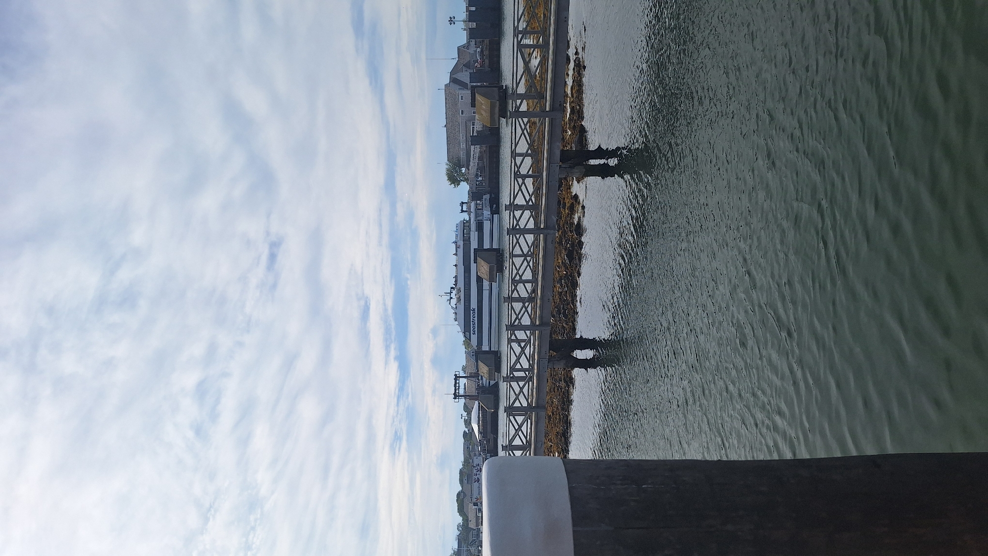In Nantucket, Massachusetts, a ferry named "SEA STREAK" is docked at a terminal. The ferry is a modern vessel with a white hull and black accents, and several passengers are visible on its upper deck. In the foreground, a wooden pedestrian bridge with a criss-cross railing extends over the water, connecting to a small, rocky island. The water is dark and slightly rippled, reflecting the overcast sky. Beyond the terminal, buildings with gray roofs and white siding are visible, suggesting a coastal town setting. The sky is filled with a mix of white and gray clouds, with patches of blue peeking through.