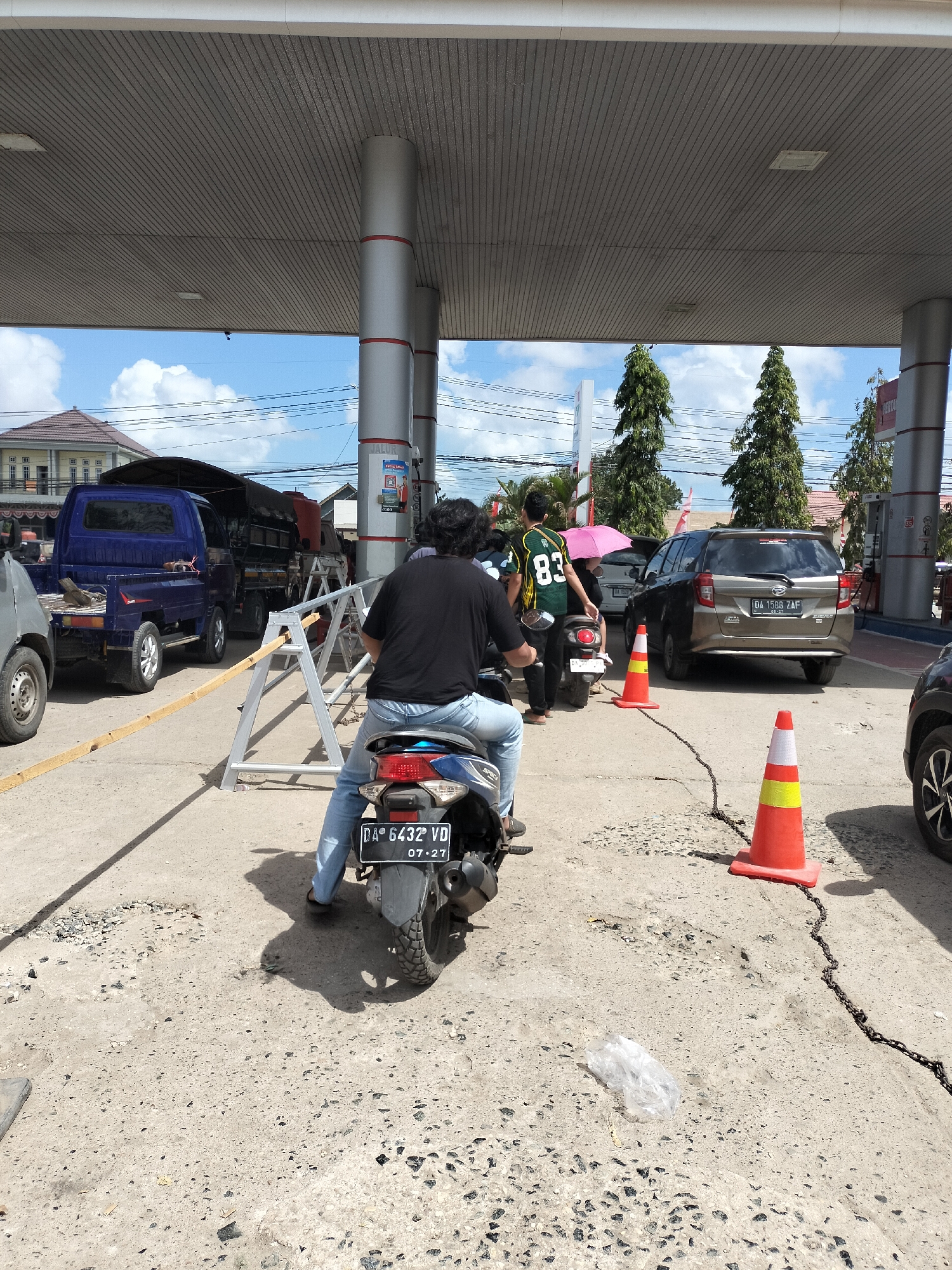 The image shows a scene at a gas station in Angsana, Indonesia. In the foreground, a person on a motorcycle, wearing blue jeans and a black t-shirt, is facing away from the camera. The motorcycle's license plate reads "DA 6432 VD" and "07-27". In the middle ground, there are several vehicles, including a blue pickup truck, a black truck with a red tank, a brown station wagon, and another motorcycle with a person holding a pink umbrella. Orange traffic cones are placed on the ground, with a chain connecting them. In the background, there are buildings, trees, and a cloudy sky. The ground appears to be made of concrete and gravel.