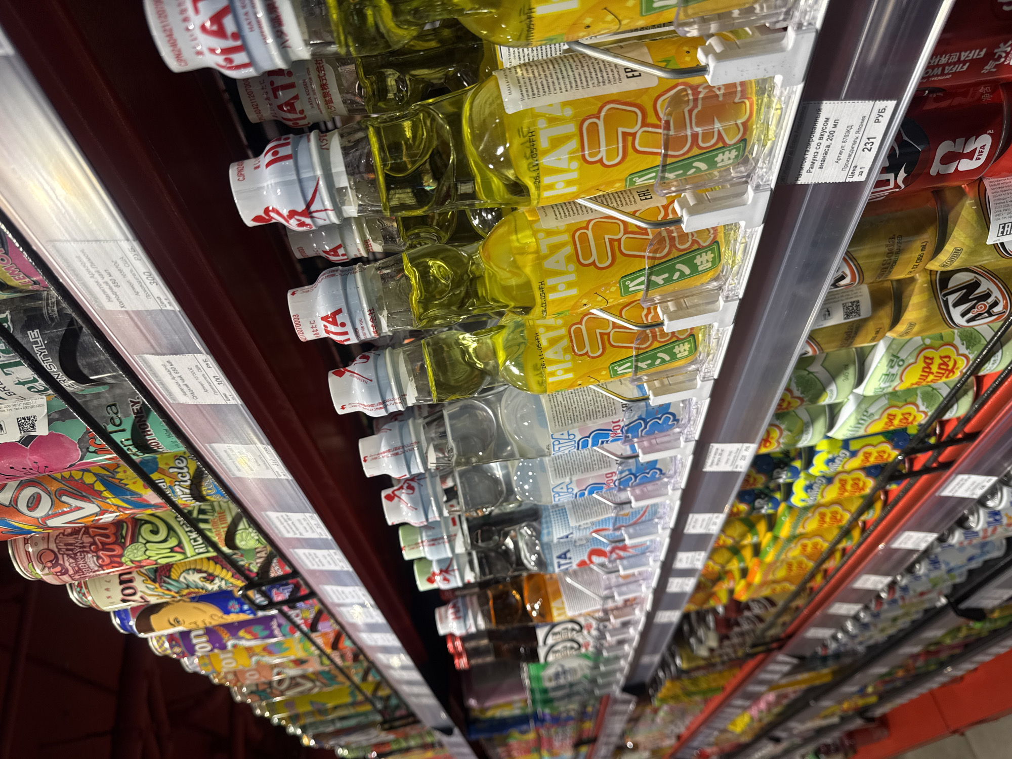 The image depicts a shelf in a store, likely in Saint Petersburg, Russia, given the product labels in Russian. The shelf is stocked with various beverages, including "Arizona" iced teas, "Ramune" soda, and "Hata" drinks. The products are arranged in rows, with price tags displayed on the shelf edges indicating the cost of each item. The overall perspective is a worm's-eye view, looking up along the shelf. The environment is a retail setting, possibly a supermarket or convenience store.