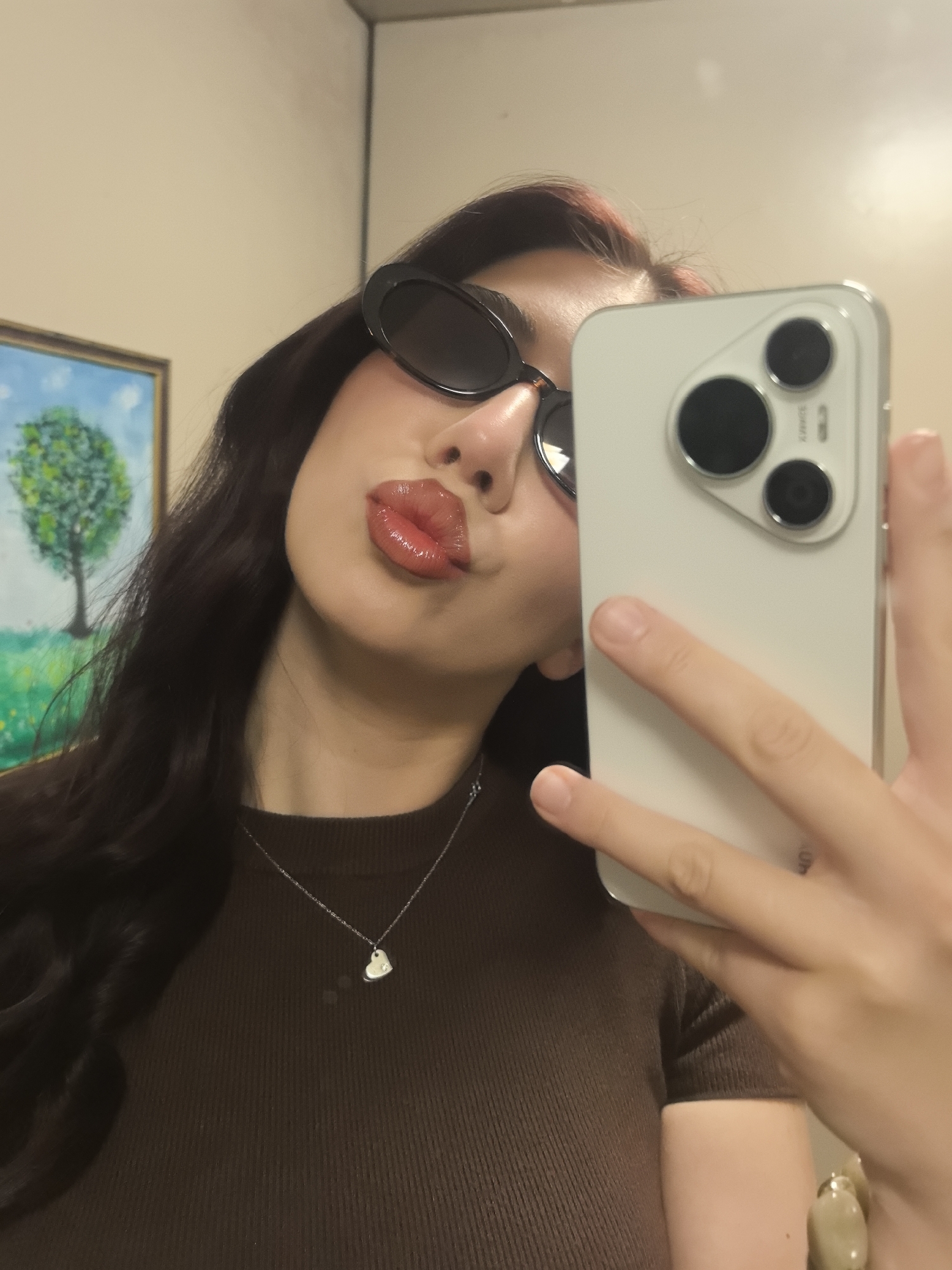 A person is taking a selfie with a white smartphone. The person is wearing black sunglasses, a brown ribbed shirt, and a silver necklace with a heart pendant. The person is pursing their lips. A painting of a tree is visible in the background. The location is Unknown, Unknown.