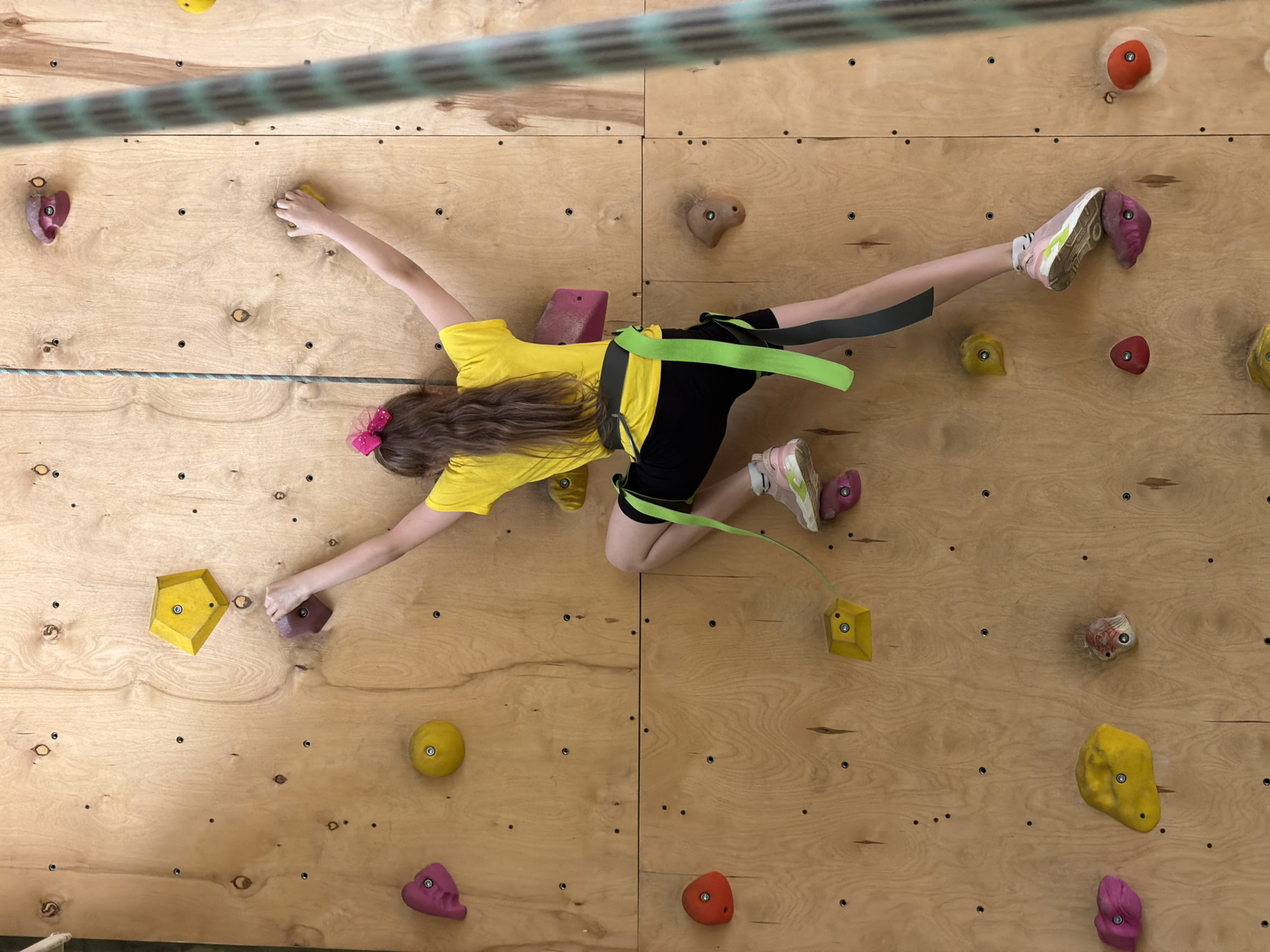 A young girl is climbing a rock wall. She is wearing a yellow t-shirt, black shorts, and pink and white sneakers. She has long brown hair and a pink bow in her hair. She is wearing a green climbing harness. She is holding onto various colored climbing holds on the wall. The wall is made of light-colored wood with multiple drilled holes. A thick rope is hanging above the girl. The location is possibly Izhevsk, Russia.
