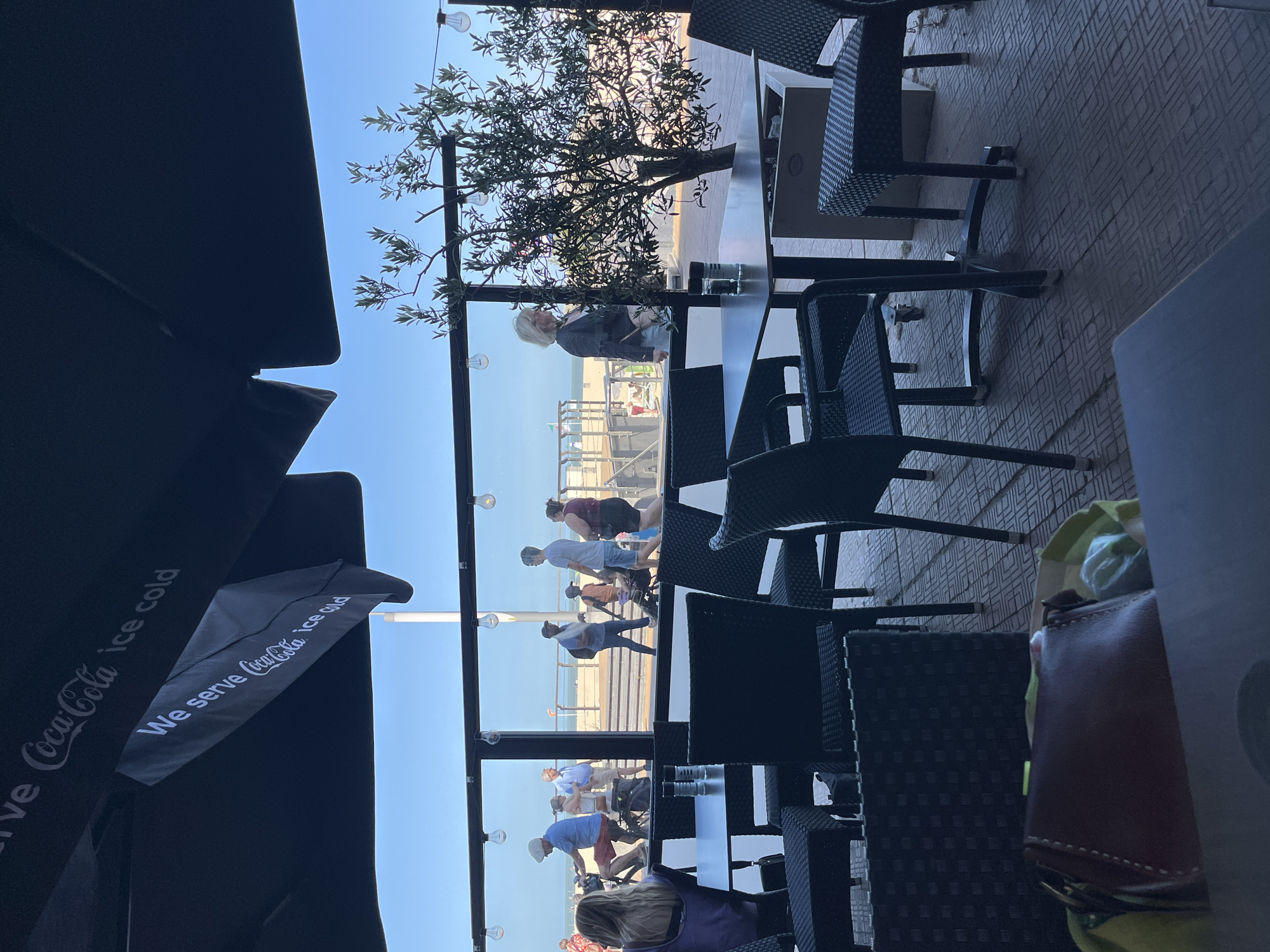The image captures an outdoor seating area of a cafe or restaurant in Ostend, Belgium, likely on the beachfront promenade. In the foreground, several tables with black wicker chairs are visible. One table has salt and pepper shakers. A dark-colored purse is placed on the floor near a table. Beyond the seating area, through a glass partition, a beach scene unfolds. People are walking along the promenade, some on bicycles, while others are strolling. The sea and sky form the background. Overhead, a large black umbrella shades the seating area, with "We serve Coca-Cola ice cold" printed on it. A small tree with sparse leaves grows near the partition, and string lights are visible above. The overall impression is a sunny day at a seaside establishment.