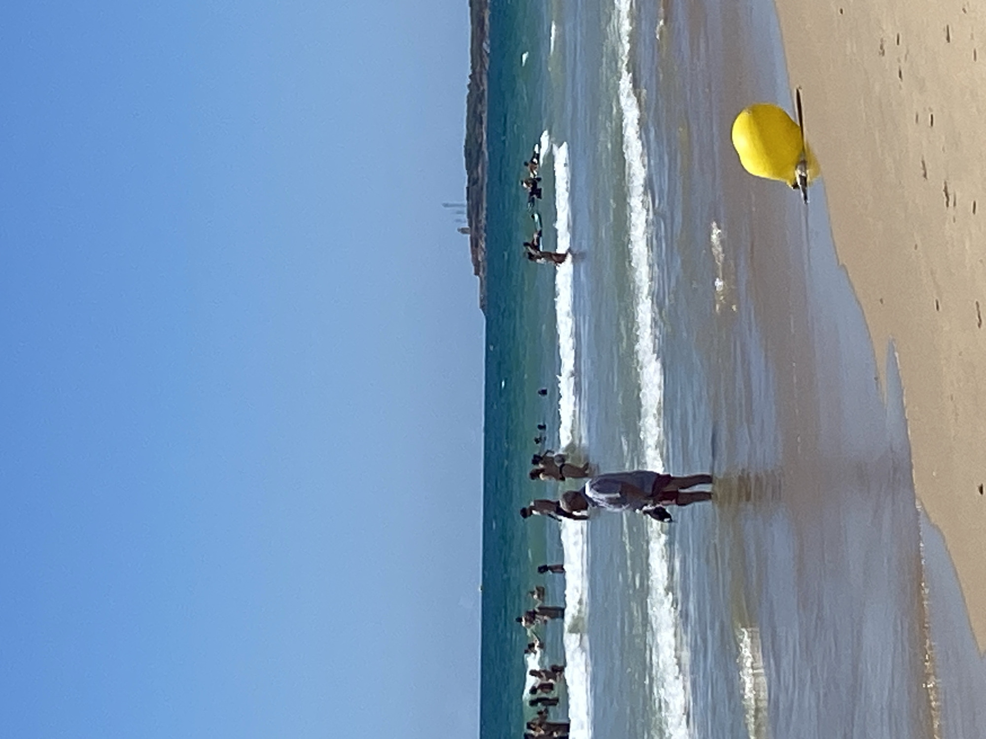 This image depicts a beach scene in Conil de la Frontera, Spain. The foreground is dominated by a sandy beach with a yellow buoy partially submerged at the water's edge. In the middle ground, several people are visible wading and swimming in the ocean, with waves breaking on the shore. A man wearing a light-colored shirt and red shorts stands in the shallow water, holding a dark object. The background shows more people in the sea and a distant coastline with structures, possibly lighthouses, against a clear blue sky. The water is a vibrant turquoise, suggesting a sunny day.