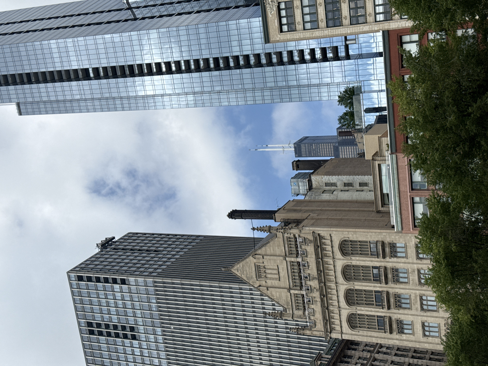 This is a photograph of an urban skyline, likely in Chicago given the reference context. The image is oriented vertically. Several tall buildings are visible, featuring a mix of modern glass-facade structures and older, more ornate architecture.
In the foreground, a large, dark-colored building with a grid of windows dominates the lower left portion of the frame. To its right, a tan-colored building with arched windows and detailed stonework stands out, topped by a dark smokestack. In the background, a very tall, sleek glass skyscraper reflects the sky and clouds. Other buildings of varying heights and architectural styles are interspersed throughout the scene, including a red brick building and a dark gray building with a pointed top. Lush green trees are visible on the right side, framing the architecture.
The sky is partly cloudy, with patches of blue sky visible, suggesting it's daytime. The lighting indicates it could be morning or afternoon. There are no people or specific activities depicted, making it a static architectural scene. The image's composition, with the buildings filling the frame and the upward tilt, emphasizes the verticality and scale of the urban environment. There is no visible text in the image.