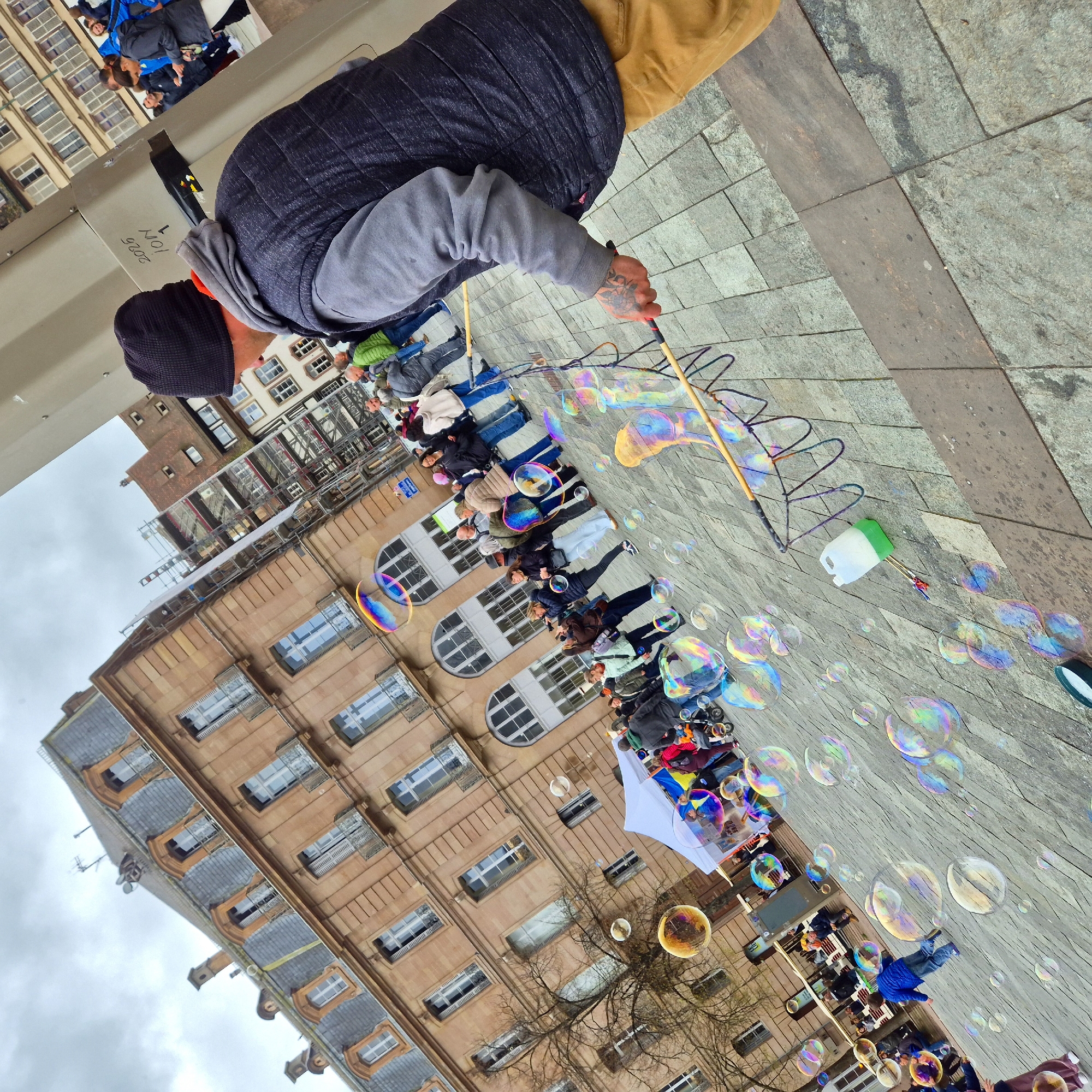 The image shows a street scene in Strasbourg, France, with a man creating large soap bubbles. The man is wearing a black beanie, a gray sweatshirt, a dark blue quilted vest, and khaki pants. He is holding a bubble wand made of wood and rope and is surrounded by numerous iridescent soap bubbles of varying sizes. A white plastic container with a green lid is on the ground near the bubble wand, presumably containing the bubble solution. A crowd of people, including adults and children, is watching the bubble-making activity, some of whom are capturing photos or videos with their mobile devices. The architecture of Strasbourg is visible in the background, with multi-story buildings featuring numerous windows and ornate facades. The ground is paved with stone slabs, and the sky is overcast. The overall impression is that this photo was taken during a public event or performance on a city street.