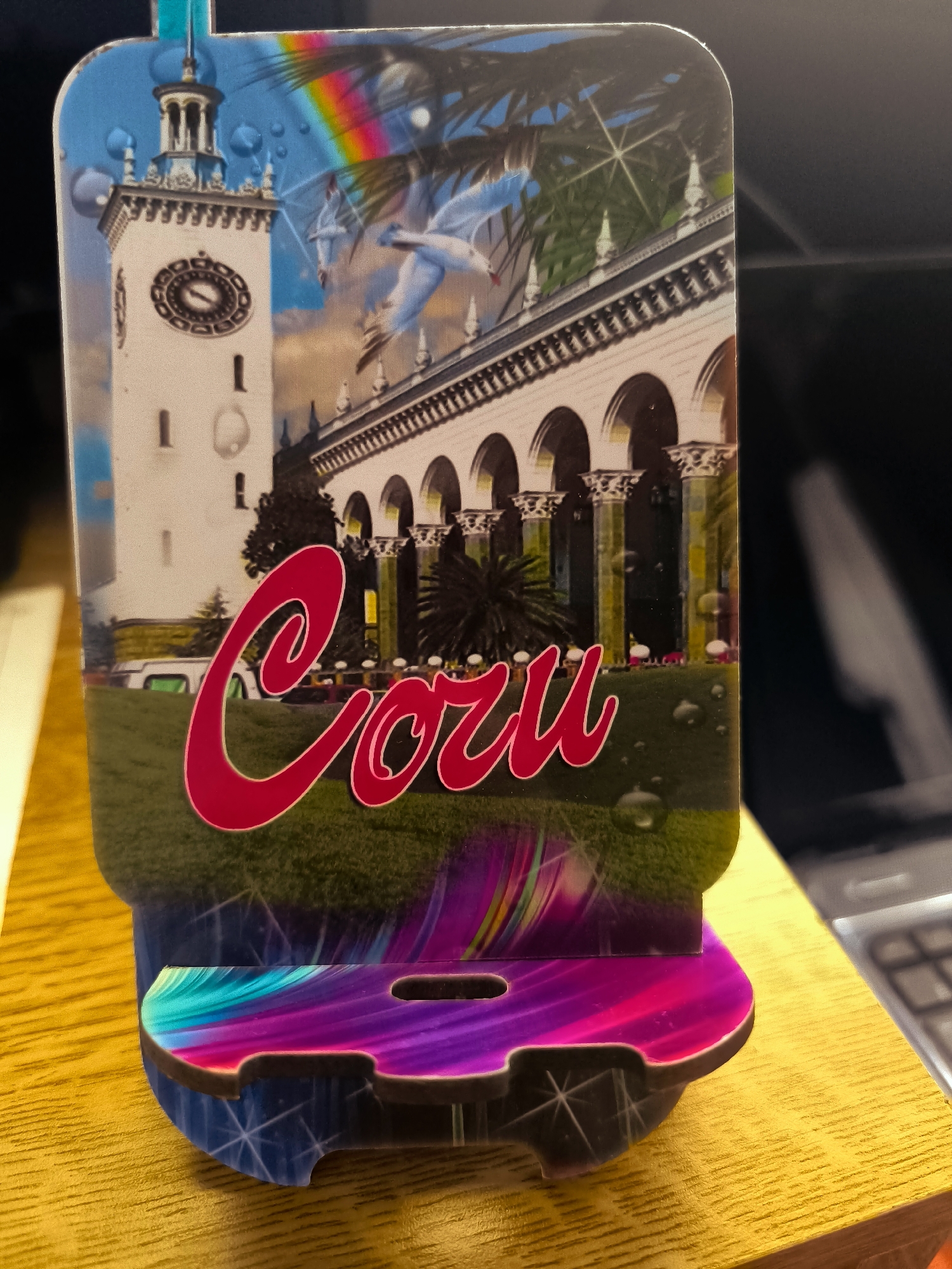 The image is a close-up, slightly angled shot of a decorative item, possibly a phone stand or a souvenir, standing on a wooden surface. The item features a colorful graphic design that includes a white building with a clock tower, arches, palm trees, a rainbow, and water droplets. In the foreground, in large, bright red cursive lettering, is the word "Coru." The base of the item has a colorful, abstract design. The wooden surface appears to be a table or desk, with a portion of a keyboard visible in the background on the right. The lighting suggests an indoor environment, possibly artificial. There is no indication of Almere, Netherlands, in the image.