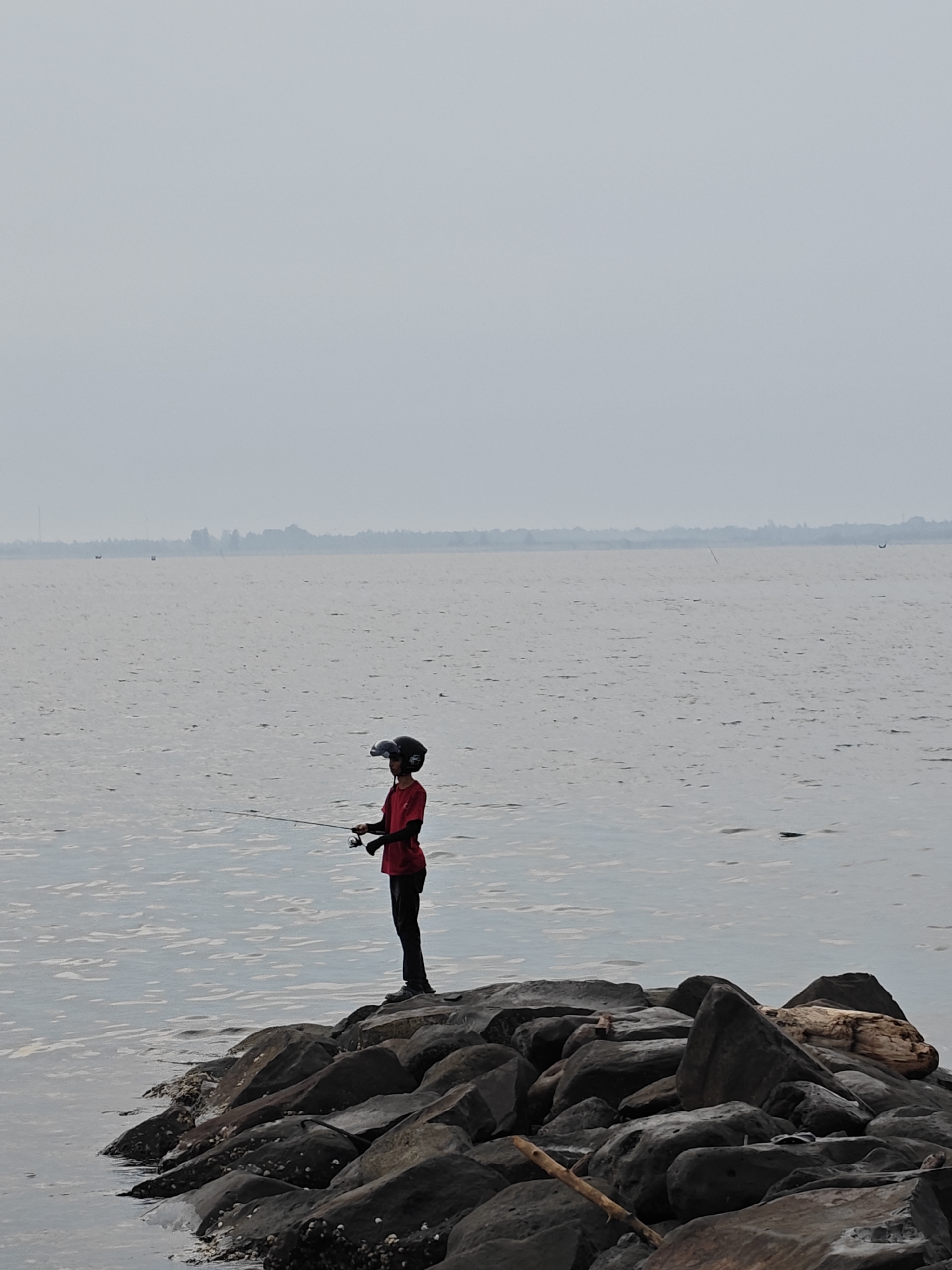 This image depicts a serene coastal scene in Lhokseumawe, Indonesia, featuring a lone individual fishing from a rocky breakwater. The main subject is a person, likely a youth, dressed in a red long-sleeved shirt, dark pants, and a dark helmet with a clear visor. They are standing on the dark, rugged rocks, holding a fishing rod cast into the water.
The scene is dominated by a wide expanse of calm, grey-blue water, suggesting a sea or a large bay. The sky above is uniformly overcast and light grey, blending seamlessly with the horizon, giving the impression of either early morning, late afternoon, or a perpetually cloudy day. The soft, diffused light creates a muted color palette throughout the image.
The foreground consists of a natural or artificial breakwater made of large, dark, weathered rocks, some of which appear wet and have small white growths, possibly barnacles or shells. Pieces of driftwood or larger logs are visible among the rocks. In the distant background, a faint, hazy landmass with indistinct trees or structures can be seen along the horizon, confirming a coastal setting. A few tiny, almost imperceptible objects, possibly boats or buoys, dot the far-off water.
The overall mood is one of peace and solitude, emphasizing the quiet activity of fishing amidst the vastness of the natural environment. There is no visible text in the image.