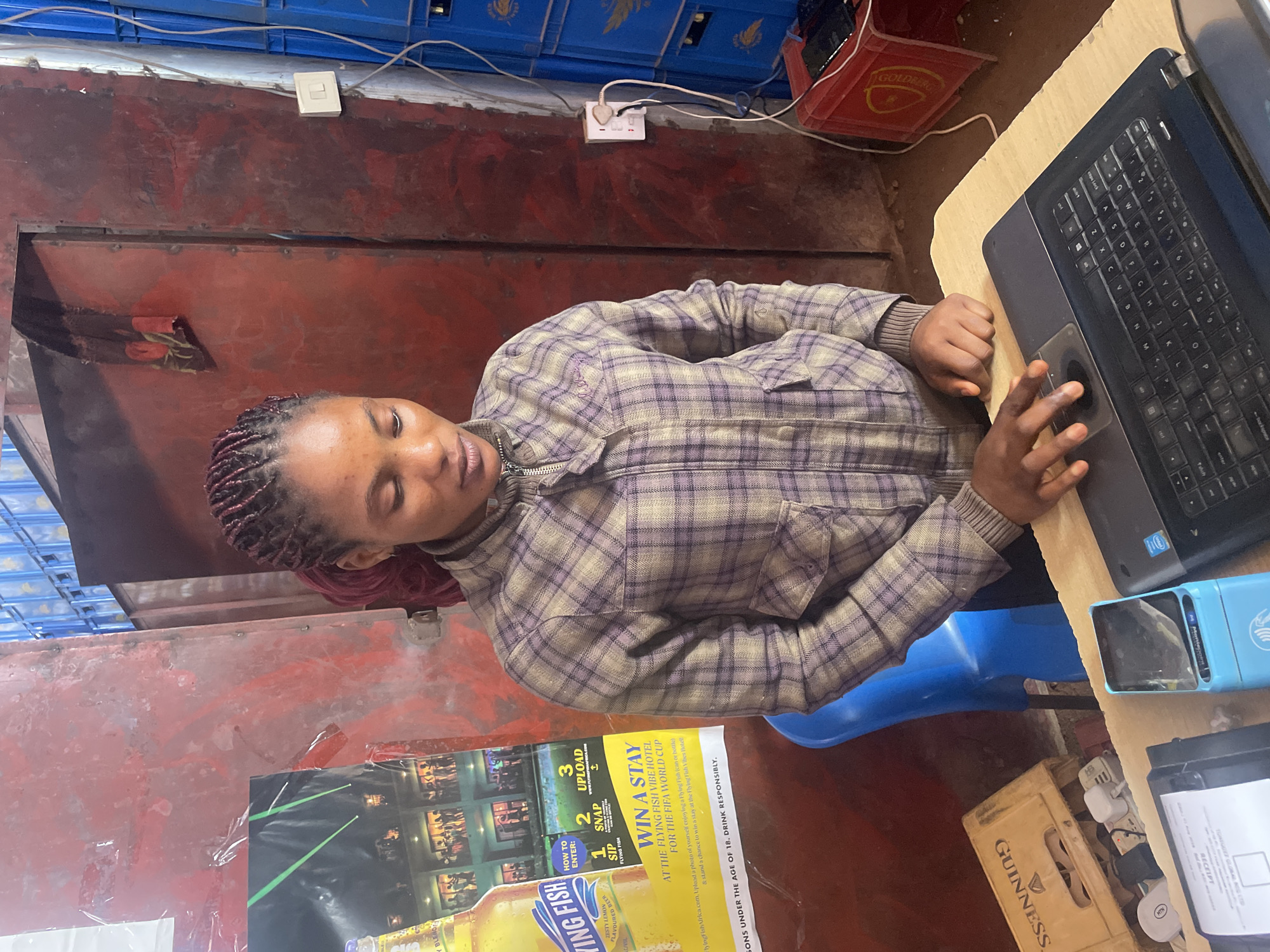 The image depicts a young woman with braided hair wearing a plaid jacket, standing behind a counter in a dimly lit, cluttered environment. She is looking down at a laptop, with her right hand resting on the trackpad. On the counter in front of her are a blue card reader, a receipt printer, and other electronic devices. Behind her, a red metal door is visible, along with stacked blue crates that appear to be filled with beverages. To her left, a poster advertising "Flying Fish" flavored beer and a Guinness beer crate are present. The overall scene suggests a small shop or bar in Gura Top, Nigeria. The lighting is somewhat harsh, with shadows contributing to the gritty atmosphere. The walls are textured and appear to be made of metal and possibly concrete.