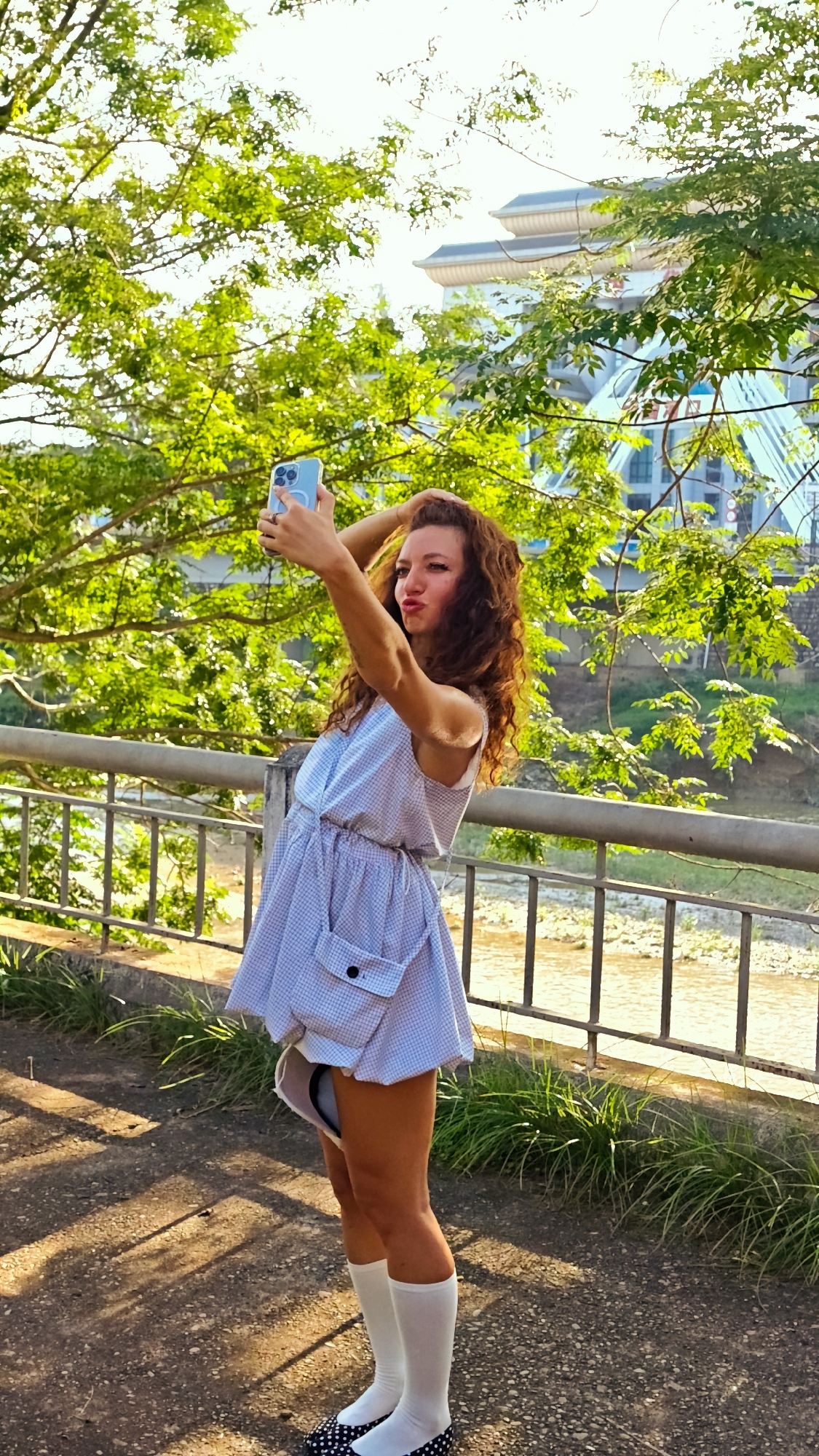 A young Caucasian woman with long, wavy auburn hair stands on a paved walkway next to a metal railing in Lao Cai Ward, Vietnam. She is dressed in a short, blue and white checkered dress with a large pocket, white knee-high socks, and black and white polka-dot shoes. The woman is holding a light blue smartphone up at arm's length, presumably taking a selfie, and has a pouting expression on her face. A bridge and a building with a tiered roof are visible through the green foliage in the background. A river is seen through the metal railing. The photograph is taken in bright daylight.