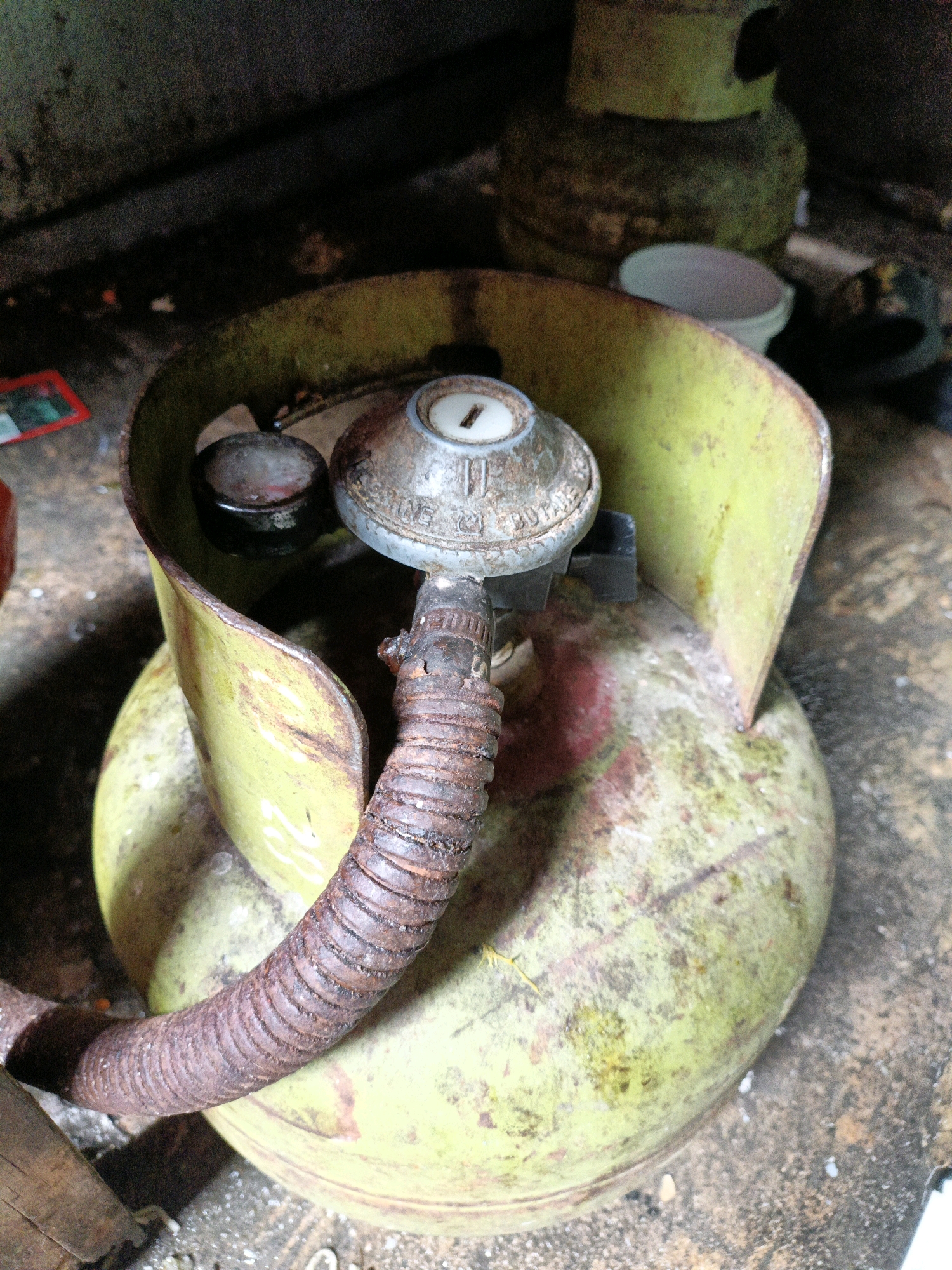 An old, green LPG cylinder is situated on a rough, concrete surface. The cylinder is heavily weathered, with dirt and rust visible. Attached to the top of the cylinder is a worn, metal regulator with a hose. The regulator appears to be made of a tarnished, light-colored metal, and the hose is a flexible, ribbed material that is also heavily rusted and appears dirty. In the background, there is a dark, cluttered environment, suggesting an outdoor or storage area. Part of another LPG cylinder, also green, is visible in the background, stacked or leaning against something. A small, white plastic cup is also present in the middle ground, to the right of the main cylinder. The overall scene conveys a sense of age and disuse. The image is an overhead shot, looking down at the cylinder.