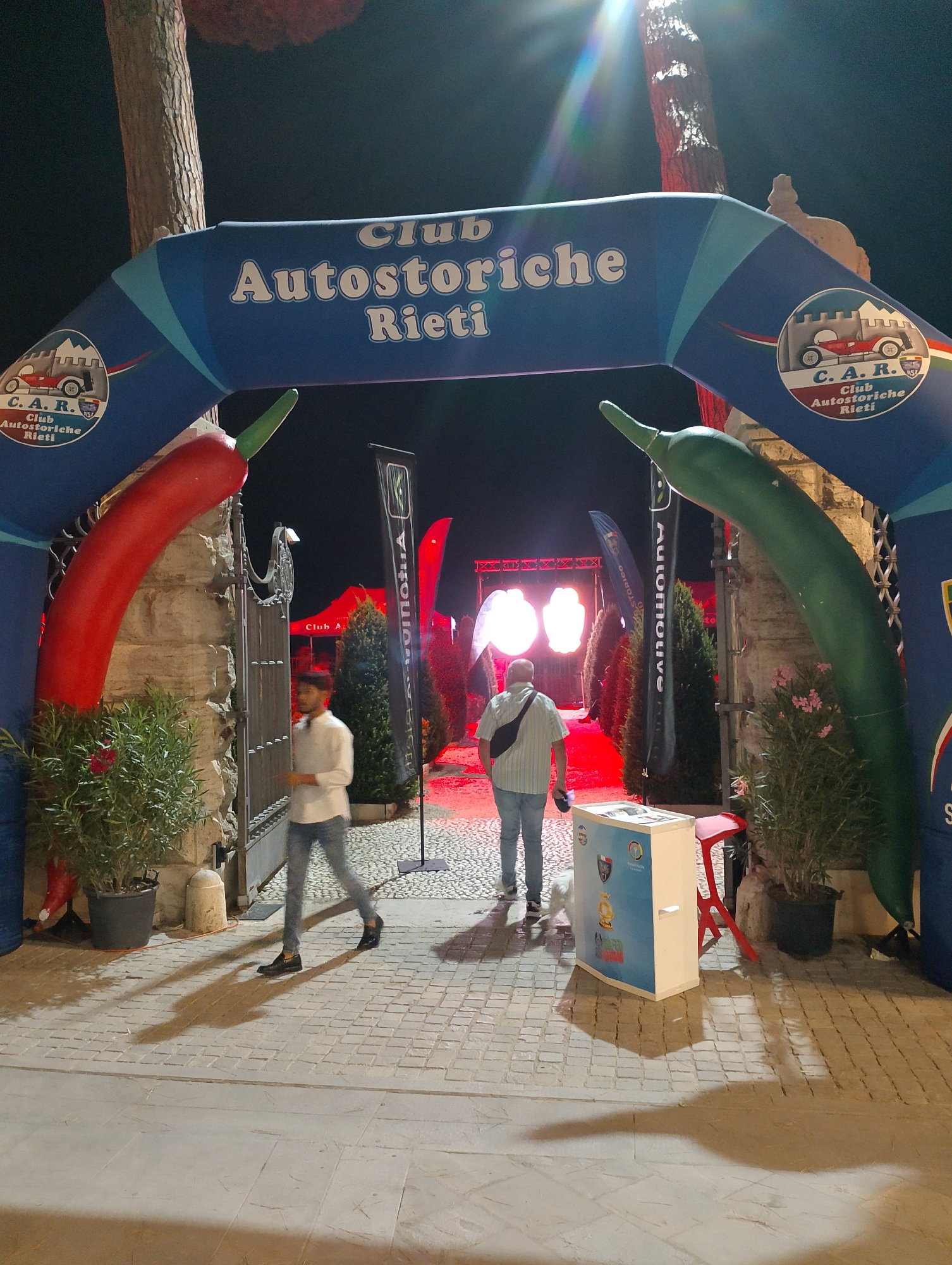 The image depicts an outdoor scene at night, possibly an event or gathering, with a prominent inflatable archway. The archway is blue with white lettering that reads "Club Autostoriche Rieti." On the left side of the arch, there's a circular logo with "C. A. R." and "Club Autostoriche Rieti" below it, featuring an illustration of a vintage car and a castle. Giant inflatable chili peppers, one red on the left and one green on the right, flank the entrance beneath the arch.
Two men are visible walking through the archway. The man on the left is wearing a white button-down shirt and blue jeans, walking towards the left of the frame. The man on the right is wearing a striped short-sleeved shirt, blue jeans, and a black fanny pack, walking towards the right of the frame.
Behind the archway, there are several black banners with white lettering, some of which read "Automotive." In the background, there are large, illuminated shapes that appear to be lights or stage props, casting a red glow. Trees and plants are visible, and the ground is paved with cobblestones. The overall atmosphere suggests a festive or commemorative event. The event is likely located in Rieti, Italy, as indicated by the signage.