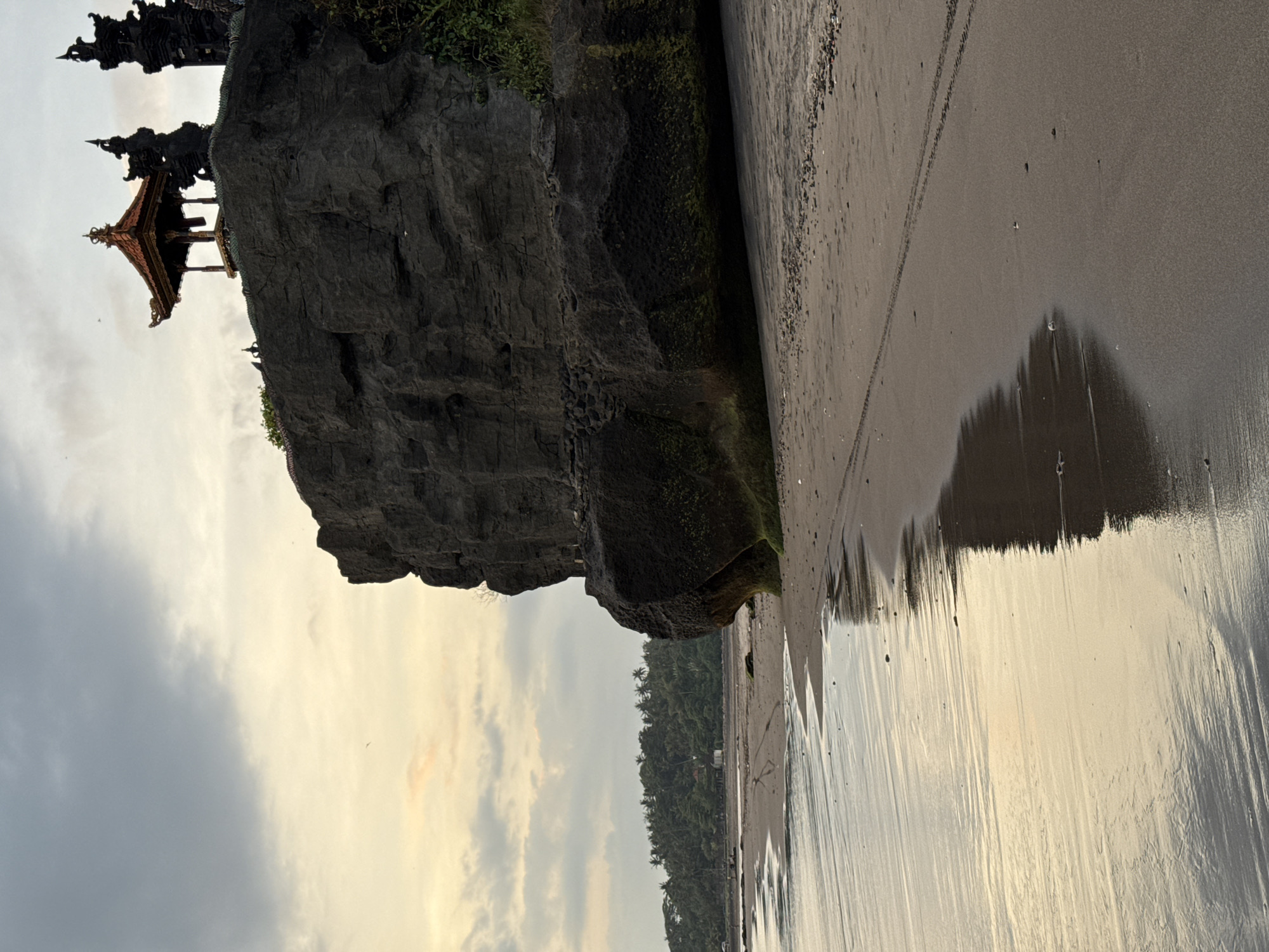 The image shows a rocky cliff with a temple structure perched atop it, located on a beach. The beach has dark sand, wet from the tide, reflecting the sky. The sky is overcast with hints of orange near the horizon. The scene includes a view of trees in the background. The image is rotated. Location: Unknown, Unknown.
