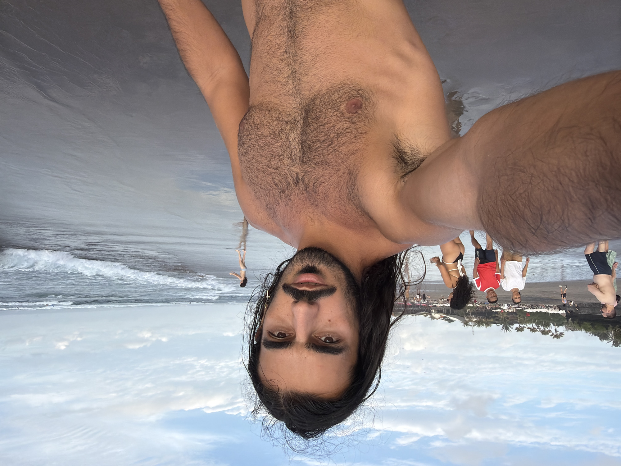 A selfie is taken by a shirtless man with long dark hair and a mustache, standing on a beach in North Kuta, Indonesia. The man is looking directly at the camera with a neutral expression. He has a prominent chest and abdomen covered in dark hair, and arm hair is visible.
In the background, several people are walking on the wet sand near the water's edge. To the left, a man in dark shorts and a lighter colored top is walking away from the camera. Further left, a couple is visible; the woman is in a white top and the man in a red top, both wearing shorts. A woman in a black bikini is seen in the middle ground, facing away from the camera. On the far right, a small figure of a person is standing near the water.
The ocean waves are breaking on the shore, and the water reflects the cloudy sky. The beach itself appears dark and wet. Palm trees and some structures are visible in the distance along the coastline, characteristic of a tropical beach resort area. The overall lighting suggests it is either early morning or late afternoon.