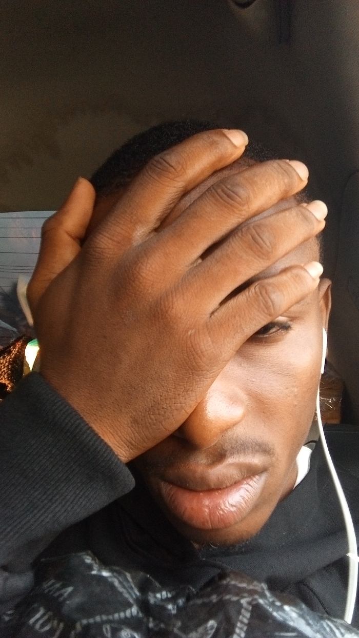 A young Black man with close-cropped hair covers part of his face with his right hand. A single eye peers out from behind his fingers. He has full lips and a neatly trimmed mustache. He wears a black hoodie, and white wired earbuds are visible. The background is blurry, possibly a car interior. The man appears pensive or possibly shielding his eyes from light.