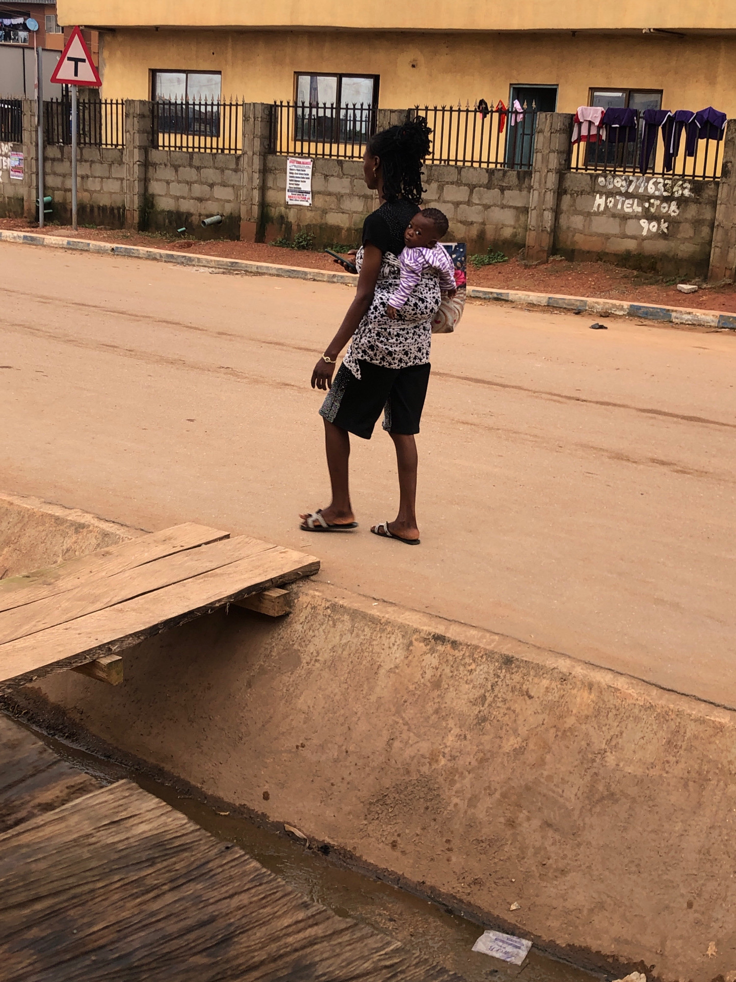 The image shows a woman carrying a baby on her back, walking on a dusty, paved road in Lagos, Nigeria. The woman is wearing a black top with a white patterned overlay and black shorts. The baby is wrapped in a striped blanket. In the background, there is a yellow building with windows and a fence. Clothes are hung on the fence to dry. A sign on the fence has text that appears to read "HOTEL JOB 90K". To the left of the image, a triangular traffic sign with a "T" symbol is visible. A wooden plank crosses a narrow, dirty ditch in the foreground. The overall scene depicts a typical street view in a Nigerian city.