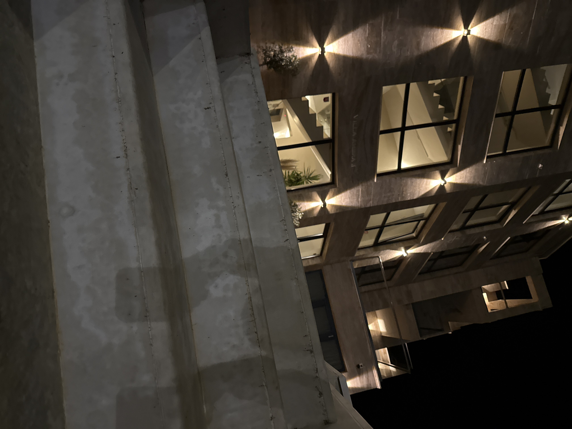 The image shows a modern building exterior at night, likely in Tyulenovo, Bulgaria. The building has a textured facade, large windows, and is illuminated by exterior wall lights. In the foreground, there are concrete steps or a tiered walkway. The steps are weathered and appear to be made of a rough-textured material. The building has multiple stories, with balconies or terraces visible on the upper levels. The overall impression is one of contemporary architecture set against a dark night sky.