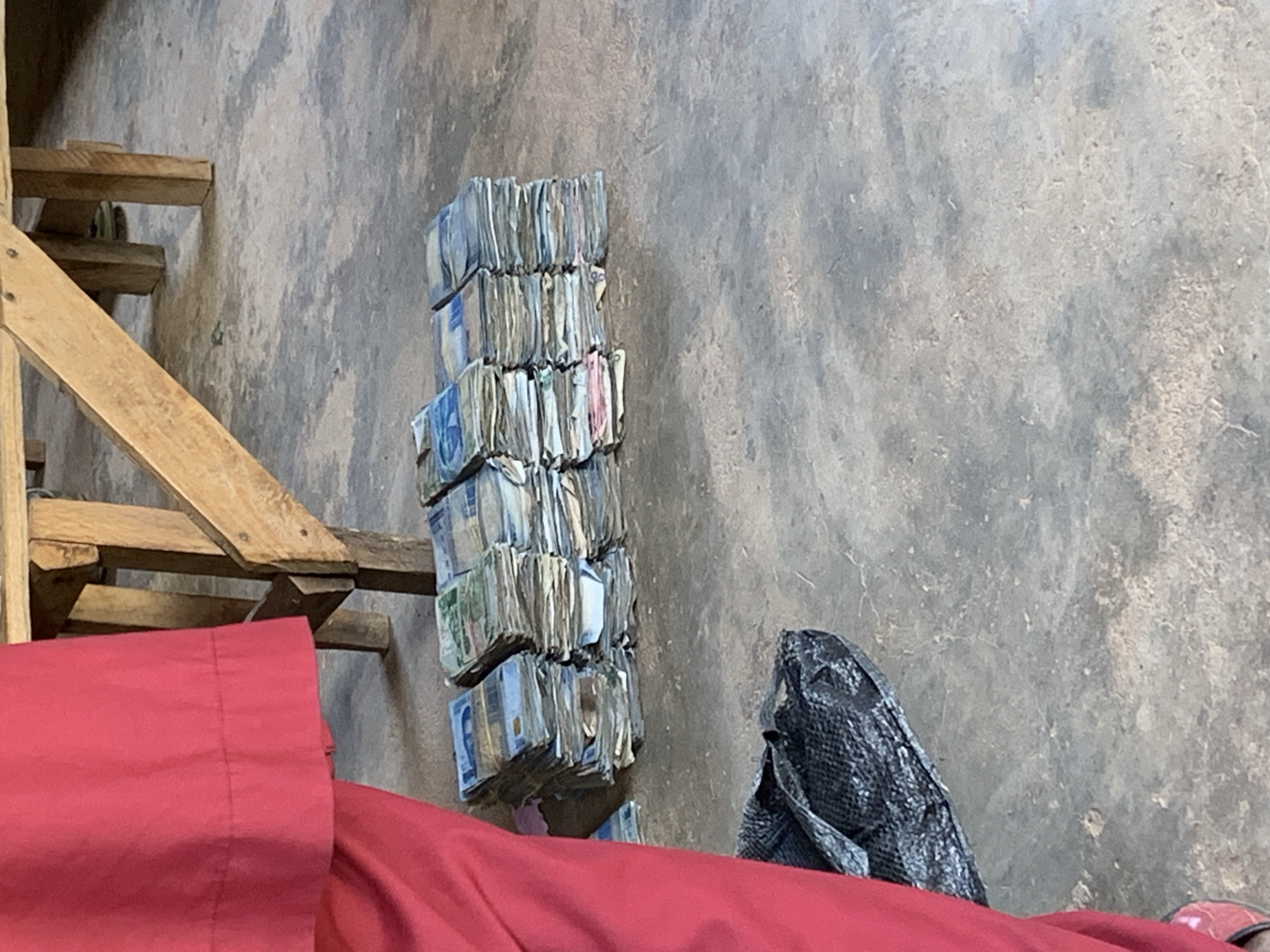 A vertical stack of paper currency sits on the floor next to a black bag, in Jos, Nigeria. A section of a wooden ladder is visible in the upper left of the image. A red cloth is in the foreground. The floor is concrete or similar material.