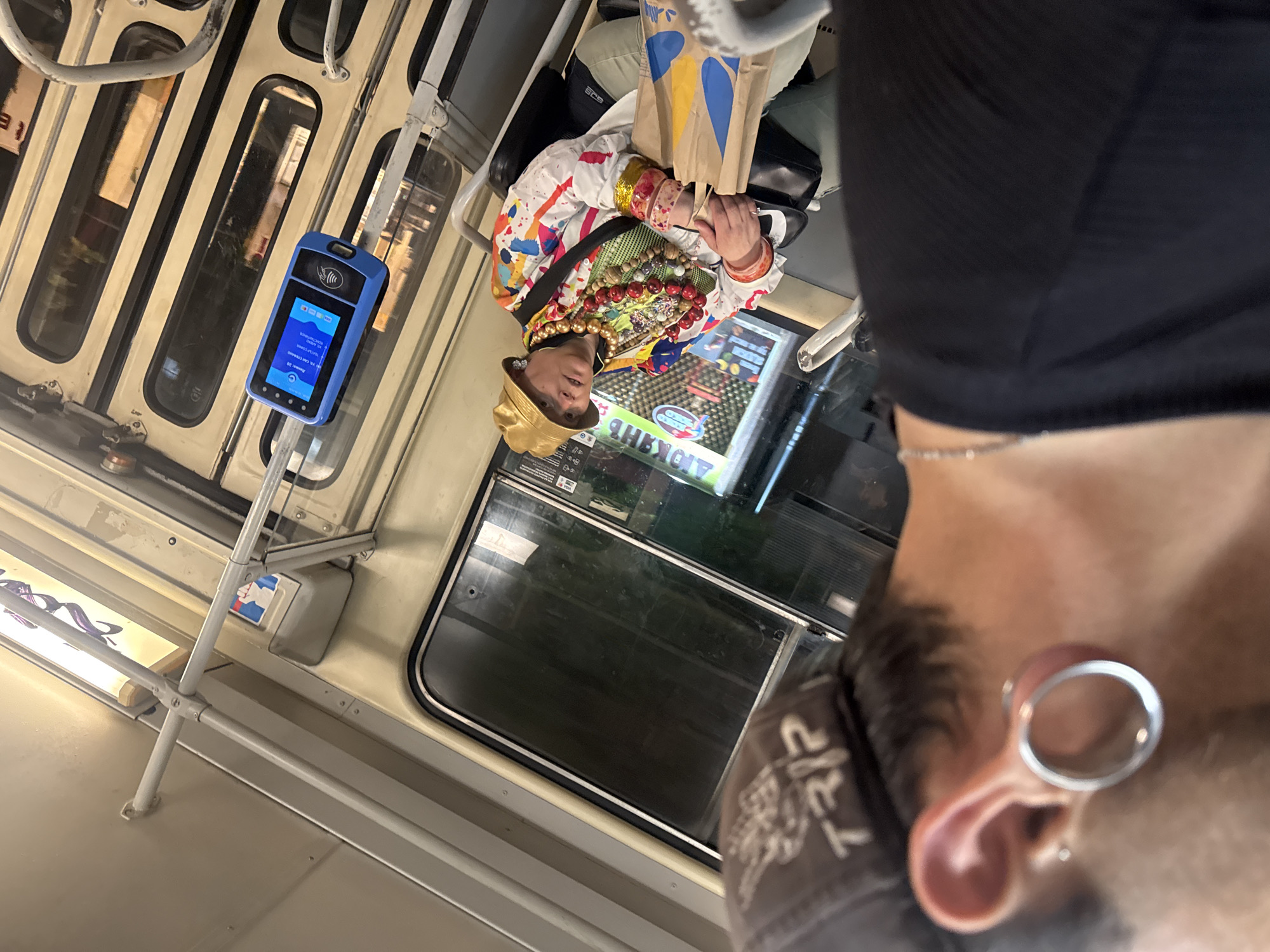 The image depicts the interior of a public transportation vehicle, likely a tram or metro car, given the interior design and the presence of a fare validator. A person, viewed from behind and the side, is in the foreground. This individual has a large circular gauge in their left ear and wears a dark baseball cap. In the mid-ground, another person is seated. This individual wears a gold bucket hat, an ornate necklace, a brightly colored, multi-patterned jacket, and several bracelets. They are holding a brown paper bag with blue and yellow markings. A blue electronic fare validator with a screen showing text and icons is mounted on a pole to the right of the seated person. The vehicle's interior features cream-colored walls, metal poles, and windows showing a blurred urban or station environment outside. The visible signage on the fare validator and within the vehicle suggests it may be a public transport system in Sofia, Bulgaria, as indicated by Cyrillic text and familiar transport icons often used in European transit systems.