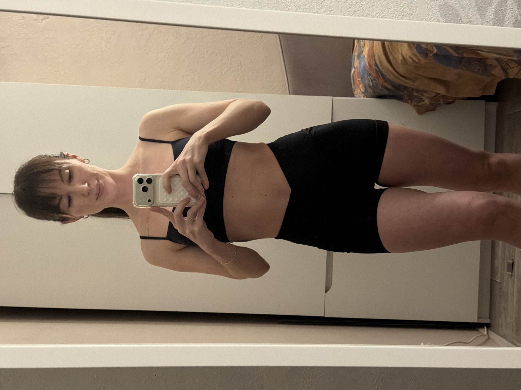 A young woman is captured in a full-body mirror selfie taken indoors in what appears to be an apartment in Perm, Russia. She is wearing a two-piece black athletic outfit, consisting of a sports bra or bikini top with thin straps and matching black shorts. Her dark hair features bangs, and she has a slender physique, with a thin bracelet visible on her left wrist.
She holds a light-colored smartphone with a textured case at chest level to take the photo, looking down towards the screen with a neutral or slightly smiling expression. The background visible in the mirror reflection includes a large white wardrobe or wall behind her on the left. To the right, a bed is partially visible, adorned with a patterned duvet or blanket in shades of brown, blue, and yellow. The floor reflected in the mirror appears to be a light-colored tile or laminate. The mirror itself has a clean white frame. The indoor lighting suggests the photo was taken during the day or evening. No text is visible in the image.