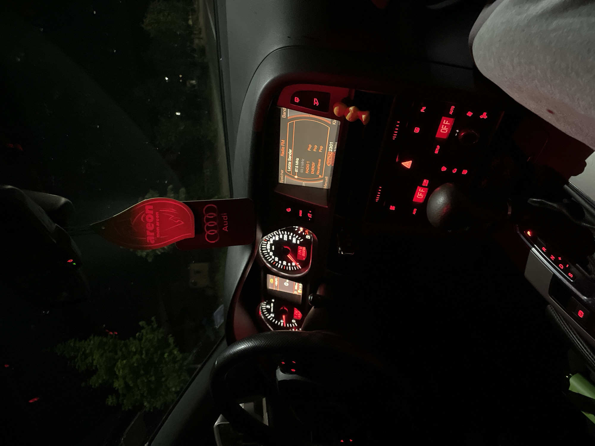 The image depicts the interior of an Audi vehicle at night in Zlatograd, Bulgaria, viewed from a low angle towards the windshield and dashboard. The car's internal lighting illuminates the dashboard, center console, and infotainment system in red. The instrument cluster shows a tachometer, speedometer, and a digital display indicating -10.0°C, zero speed, "P" for park, and an odometer reading of 253,205 km. The infotainment screen displays "Radio FM" with "Letzte Sender" (Last Stations), listing several radio stations and the time as 23:01.
A red, leaf-shaped "Areon" brand air freshener, accompanied by an "Audi" logo pendant, hangs from the rearview mirror area. A small yellow figurine is attached to the dashboard to the right of the infotainment screen. The center console features numerous red-illuminated buttons and knobs, with "OFF" and "AUTO" visible on the climate control panel, alongside the illuminated hazard light button. The gear selector displays "P R N D S" with "P" illuminated in red, indicating the vehicle is in park. A human leg, clad in grey sweatpants with a minor red mark, is visible on the right side of the frame. A green object is partially visible in the lower-left corner near the driver's side. Outside the windshield, trees and faint distant lights are visible against the dark night sky.