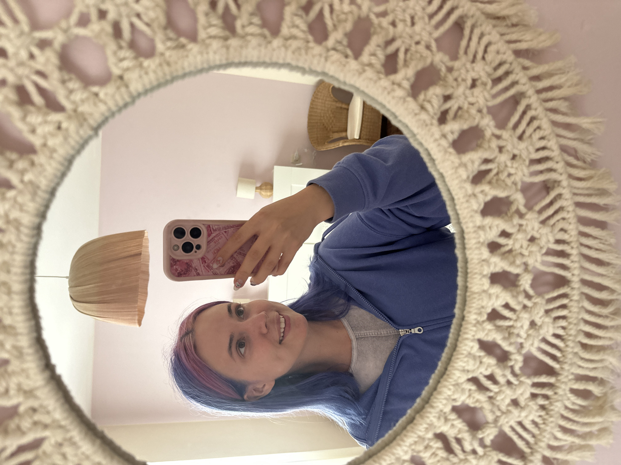 A selfie in a mirror reveals a young woman with blue and purple hair, wearing a blue zip-up hoodie and a grey top. She is holding a pink phone with a patterned case. The mirror has a macramé frame. The background, visible in the mirror, shows a pink wall, a wicker basket, a white dresser, and a light fixture. The location appears to be an interior space, possibly a bedroom. The city of Gijón/Xixón, Spain is not directly evident from the image.