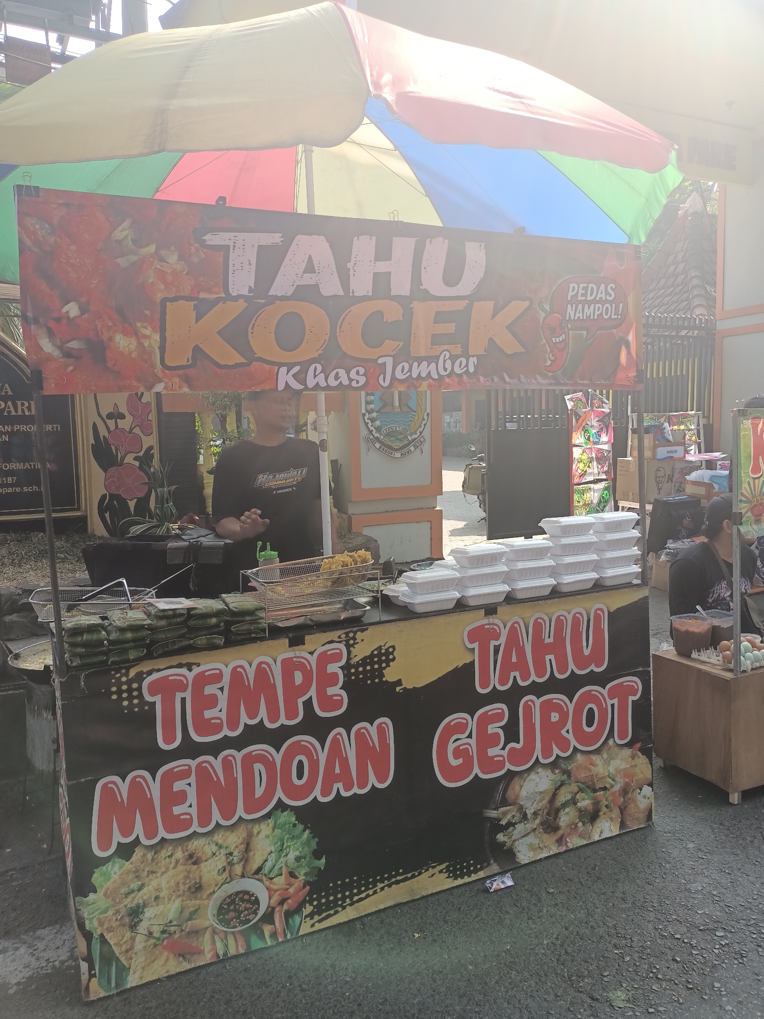 A street food stall is set up outdoors under a colorful umbrella. The stall displays a large banner with text in Indonesian that reads "TAHU KOCEK KHAS JEMBER" and "TEMPE MENDOAN TAHO GEJROT". Several stacks of white Styrofoam food containers are visible on the counter. A vendor wearing a black t-shirt stands behind the counter, interacting with customers. Various food items, including what appears to be fried tofu and tempeh, are displayed for sale. The ground is paved with asphalt, and the background shows a building with a fence and some trees. The overall scene depicts a lively street food vendor operation in the city of Pare, Indonesia.