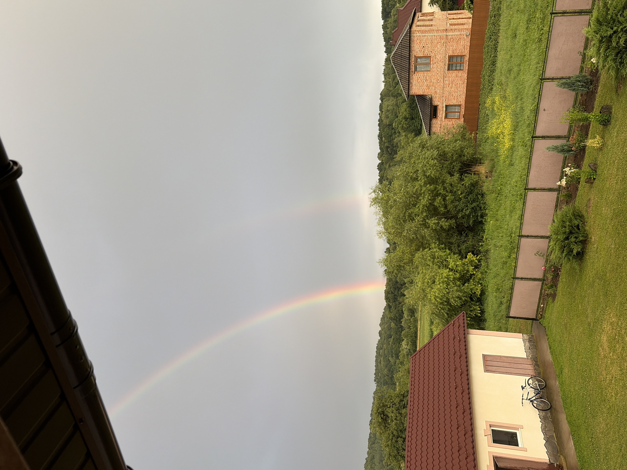 The image depicts a suburban or rural landscape with a double rainbow arching across a cloudy sky. In the foreground, a light-colored building with a burgundy roof is visible on the left, with a bicycle parked beside it. To the right, a two-story brick house with a dark roof stands behind a brown fence. The landscape is characterized by lush green grass, trees, and rolling hills in the background. The overall scene suggests a post-rain environment, indicated by the presence of the rainbow and the wet appearance of the ground and pavement. The location is identified as Hanniv, Ukraine. There are no people or animals visible in the image.