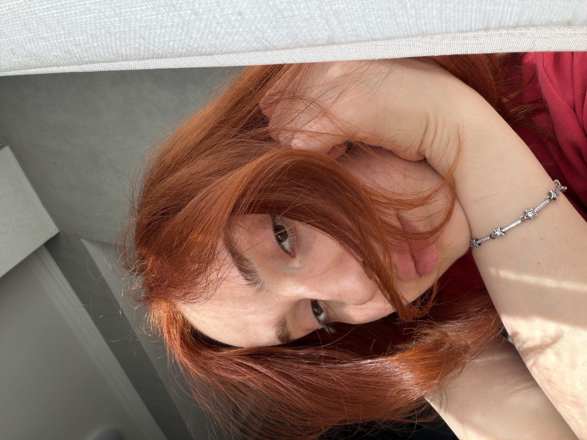 A close-up, tilted view focuses on a person with auburn hair, light skin, and brown eyes. The person is wearing a red top and a silver bracelet with small, star-shaped charms. One hand is visible near the face, seemingly supporting it. The background reveals part of a white surface, possibly a ceiling or curtain, and a gray wall.
The location is not discernible from the image. There is no way to tell if this photo was taken in Tbilisi, Georgia or anywhere else.