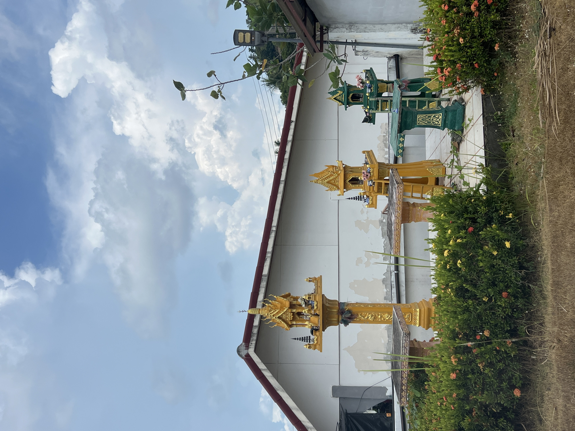 The photo shows three spirit houses placed outside a building in Baan Hua Thanon, Thailand. The houses are arranged in a line: a gold one in the foreground, another gold one in the middle, and a green one in the background. A bush with red and yellow flowers is visible between the spirit houses. The white building has a red-trimmed roof and a peeling facade. The sky is blue with cumulus clouds. A street light is visible above the building.