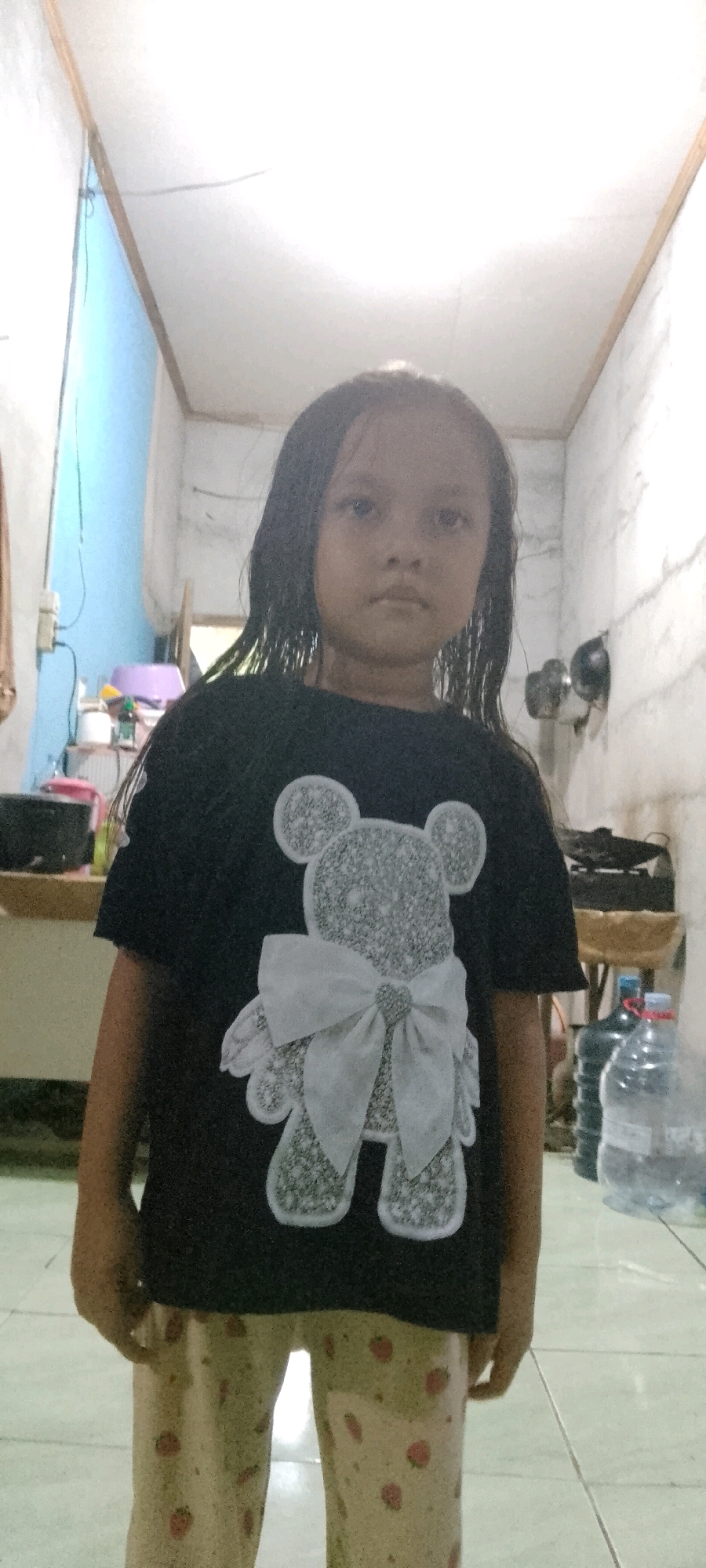 A young girl stands facing the camera in what appears to be an indoor setting. She has long, dark hair that falls around her shoulders. She is wearing a black t-shirt with a large, grey, textured teddy bear graphic on the front, complete with a white bow. Her lower body is clad in light-colored pants with a pattern of red strawberries. The background shows a room with tiled flooring and walls that appear to be painted white and a light blue. To the left, a shelf with various items, including a purple bowl and bottles, is visible. To the right, several plastic water bottles are stacked. The lighting suggests the photo was taken indoors, possibly in a home.