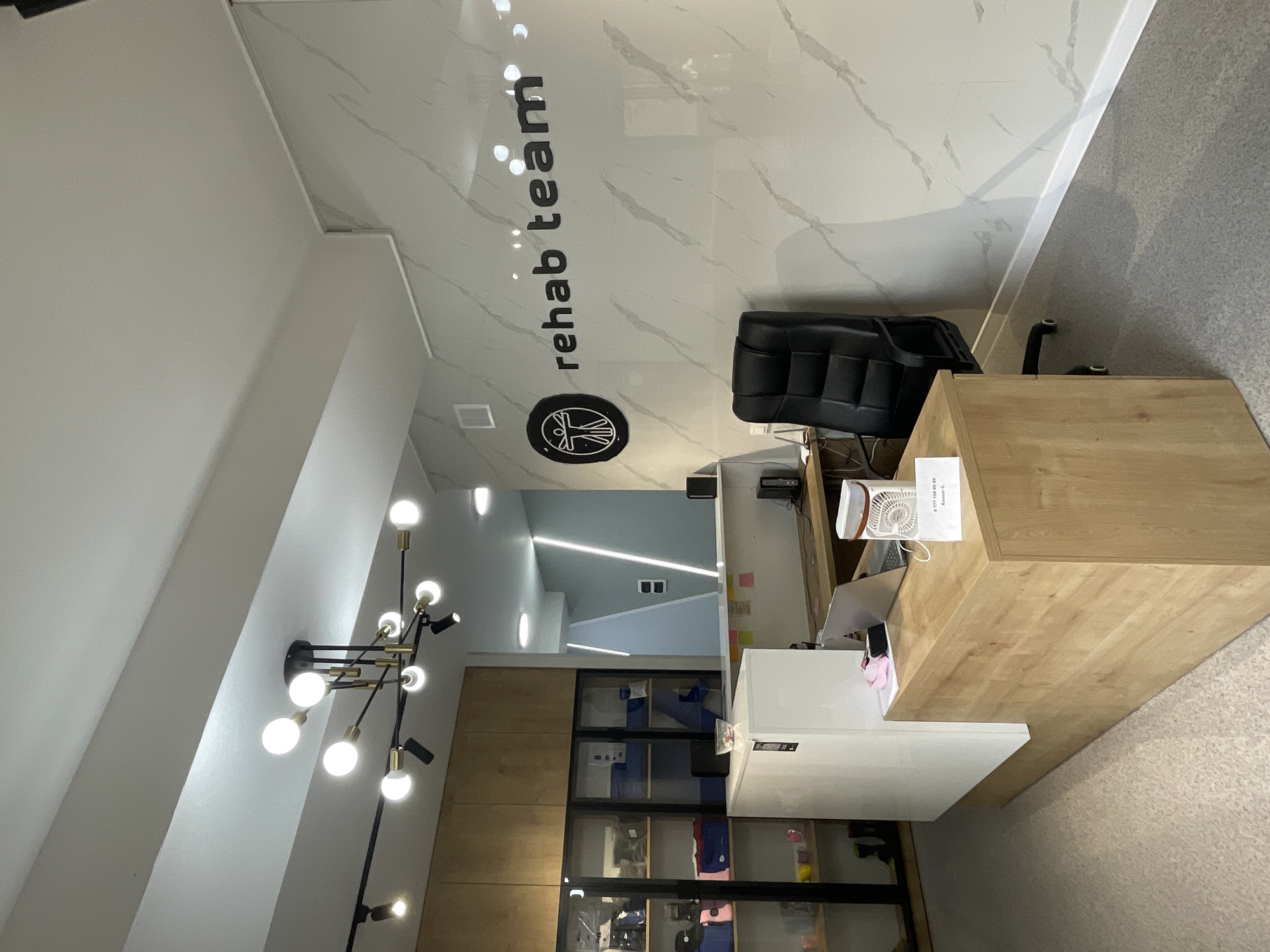 The image shows the reception area of a rehabilitation center named "rehab team" in Almaty, Kazakhstan. The reception desk is made of light-colored wood and white laminate, with a laptop, a small fan, and a sign with a phone number on it. Behind the desk, there is a black leather office chair. The wall behind the reception area is decorated with marble-effect tiles and features the "rehab team" logo. To the left of the reception desk, there are glass display cabinets with shelves containing various items, possibly rehabilitation equipment or merchandise. The overall ambiance is modern and clean, with track lighting and a contemporary chandelier hanging from the ceiling. The floor appears to be carpeted in a light grey color.