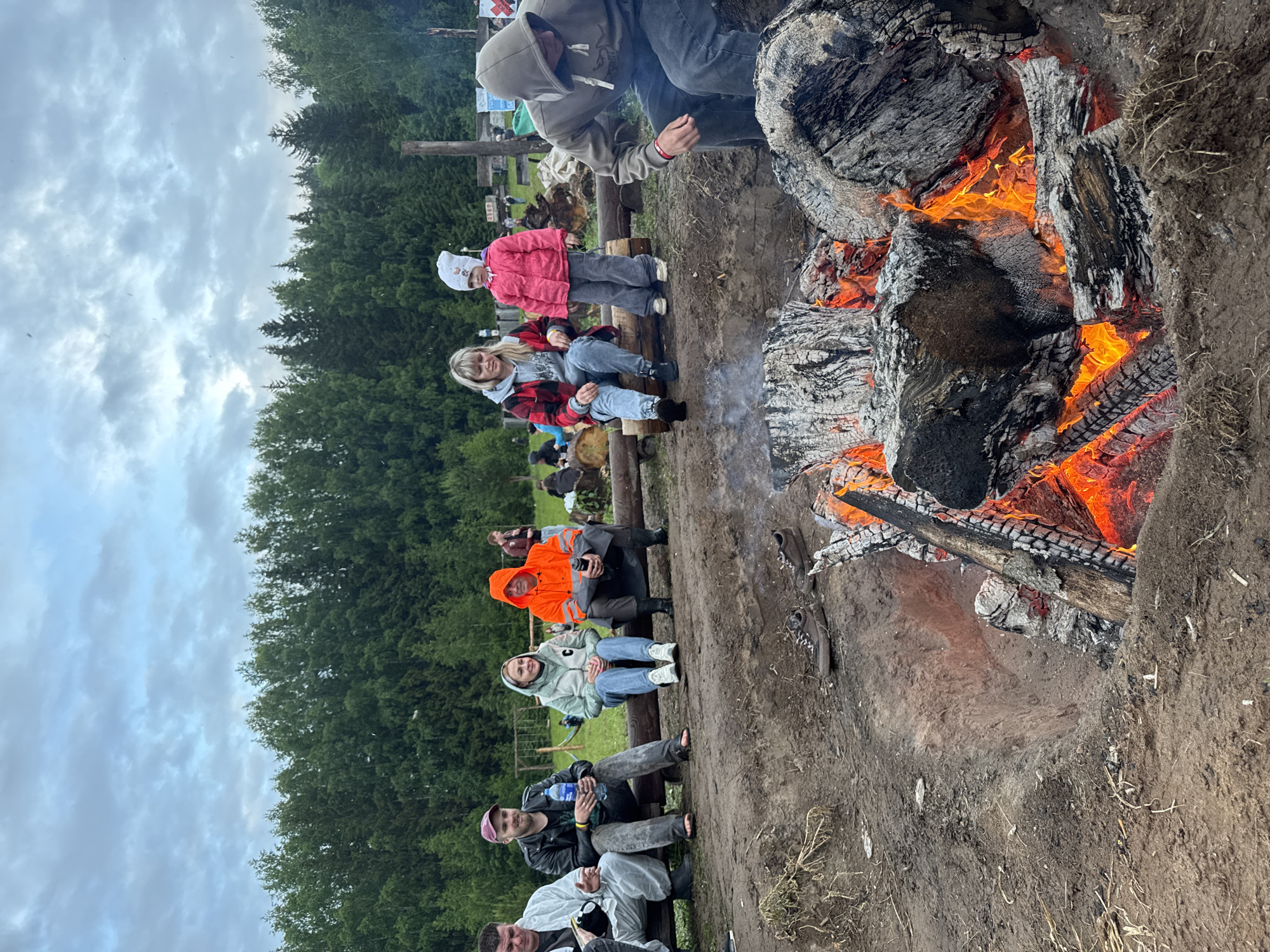 The image depicts an outdoor gathering of nine individuals around a substantial campfire in Большая Кивара, Russia. The scene is set on a dirt clearing bordered by a dense forest under an overcast sky.
In the immediate foreground, a large, excavated fire pit contains a vigorous campfire with prominent orange and red flames consuming multiple large logs, generating embers and char. Two pairs of shoes, one dark brown and one lighter brown, are resting on the dirt near the fire pit.
On the left side of the midground, five individuals are seated on a long horizontal log acting as a bench. From left to right:
1. A male in a white protective or rain suit, seated, with his left hand holding an object and his right hand slightly raised as if in conversation or gesturing.
2. A male in a black leather jacket, dark t-shirt, and ripped jeans, wearing a maroon baseball cap, holding a plastic water bottle.
3. A female in a light grey hooded jacket with a small green emblem on the chest, blue jeans, and white sneakers, looking towards her left.
4. A male in a bright orange hooded rain jacket and dark waterproof pants, holding a dark cup or bottle, looking forward.
On the right side of the midground, two individuals are seated on another horizontal log. From left to right:
1. A female in a red and black plaid jacket over a grey hoodie, blue jeans, and dark shoes, looking towards the campfire.
2. A young child, approximately 3-5 years old, in a pink jacket, grey pants, and a white knit hat, standing near the seated female and looking towards the campfire.
Closer to the right foreground, a ninth individual in a grey hooded sweatshirt and dark pants is bent forward, appearing to observe or tend to the campfire. Their face is obscured by the hood and posture.
In the background, a dense forest dominates the horizon. Several rudimentary wooden structures, resembling a swing set or playground elements, are visible in the mid-distance. Other indistinct figures are visible further back within the cleared area. The sky above is entirely covered with grey clouds.