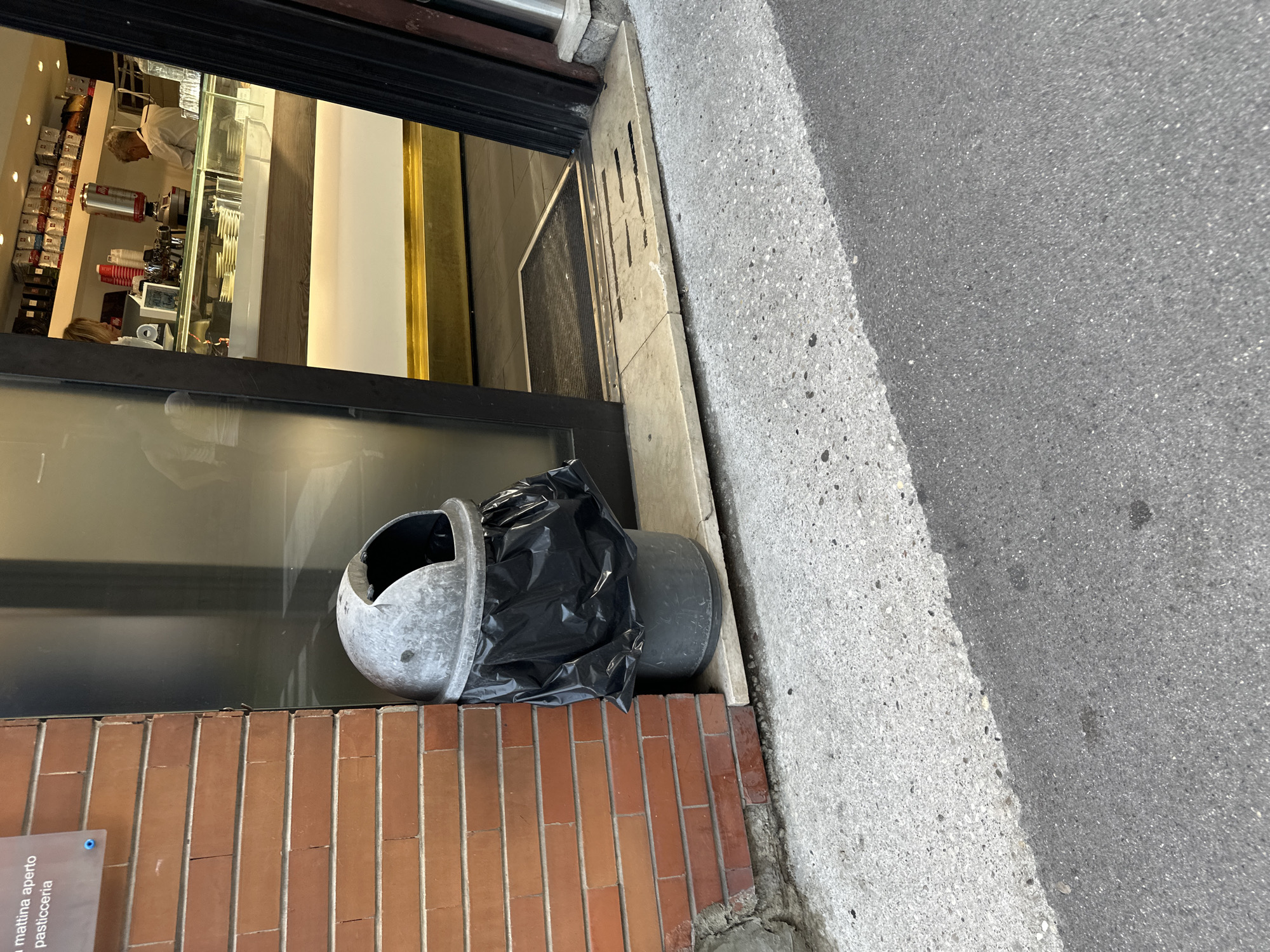 An outdoor scene shows a partially open doorway to a shop, likely a pasticceria, with a trash can in front of it. The shop interior, visible through the doorway, features a counter with a coffee machine, displays of coffee products, and two individuals, one appearing to be a customer and the other an employee. The trash can is a metal dome-shaped bin with a black plastic bag lining it, placed on a light-colored stone threshold. The building's exterior wall is made of red brick. The ground in front of the shop is paved with concrete. The image appears to have been taken during daylight hours. The shop sign above the doorway reads "La mattina aperto pasticceria," indicating it is open in the morning and is a pastry shop. While the specific location in Trieste, Italy, is not identifiable from the visual information alone, the sign's text strongly suggests a pasticceria in that city.