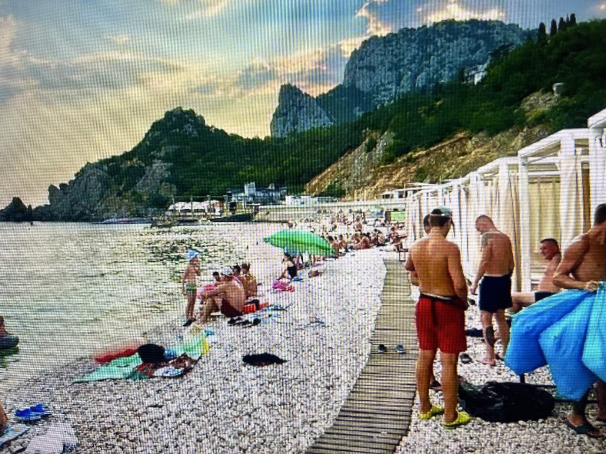 The image depicts a crowded beach scene with a pebble shore. In the foreground, several men are visible. One man in red shorts and yellow flip-flops stands facing away from the camera, while another man in dark shorts with tattoos on his leg stands nearby. To the right, a man in a light-colored shirt is partially visible. Several white cabanas with curtains are lined up along the edge of the beach. The background features a rocky, tree-covered mountainside under a cloudy sky. A body of water, likely the sea, is visible on the left side of the frame, with a few people in the water. Various beach items, such as towels and inflatables, are scattered on the pebbles. The overall scene suggests a warm, sunny day at a popular beach resort.