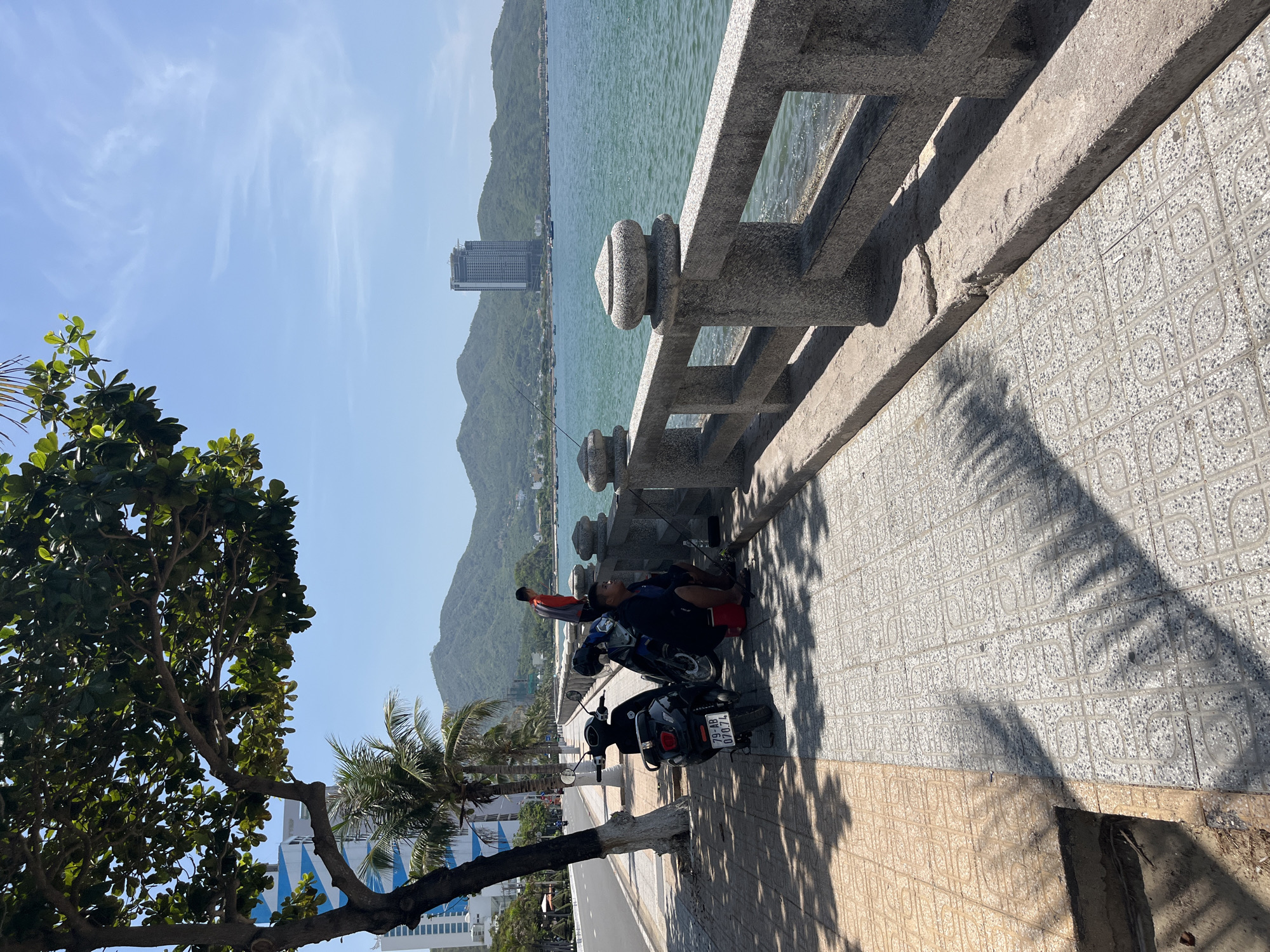 The image depicts a sunny coastal scene in Bac Nha Trang Ward, Vietnam. A wide, tree-lined promenade runs parallel to a multi-lane asphalt road on the left. The sidewalk features two distinct patterned tile sections: a tan section on the far left and a greyish-white section with a repeating geometric pattern extending towards the sea.
A large tree with dense green foliage occupies the upper left, casting prominent shadows onto the sidewalk. Behind the tree, a building with blue and white geometric patterns is partially visible. Two motorcycles are parked on the sidewalk: a black scooter with license plate "79-AB 070.74" and a partially obscured dark motorcycle.
Two men are present on the promenade. One man is seated on a red container on the grey sidewalk section, wearing a dark shirt and pants with red footwear. He is engaged in fishing, with a fishing rod extending towards the water. Nearby, a tackle box or small container is on the ground. A second man, wearing a red long-sleeved shirt and dark pants, stands further down the promenade near the stone railing, facing the water.
A grey stone railing with decorative round finials separates the promenade from the turquoise-blue sea. Numerous small fishing boats are visible on the water. In the background, a range of green, tree-covered mountains rises, with a tall, modern high-rise building prominently positioned on the distant coastline. The sky is clear blue with sparse, wispy clouds.